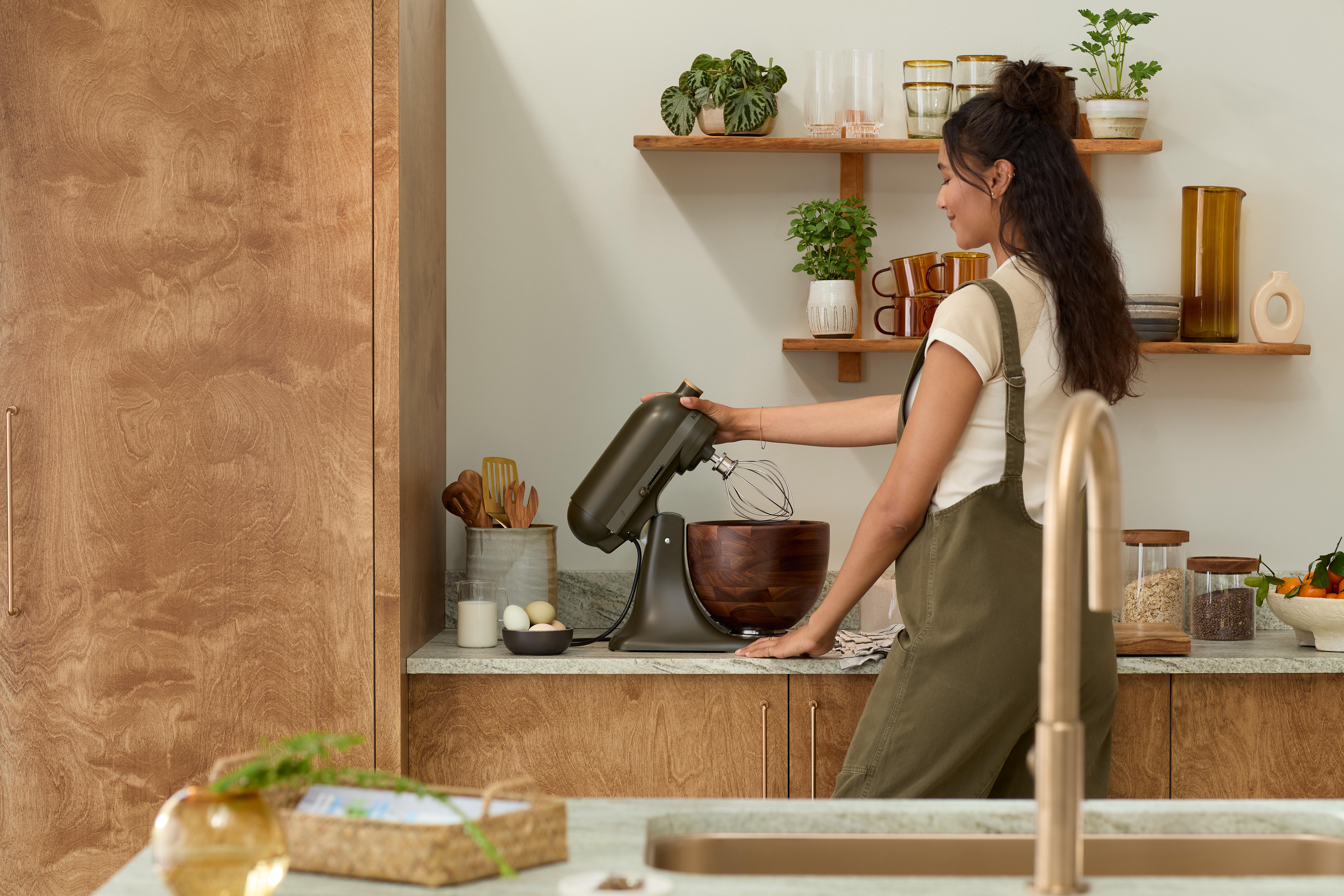 Eine Frau benutzt einen Mixer in einer Küche mit Holzschränken und Regalen. Der Mixer steht auf einer Arbeitsplatte.