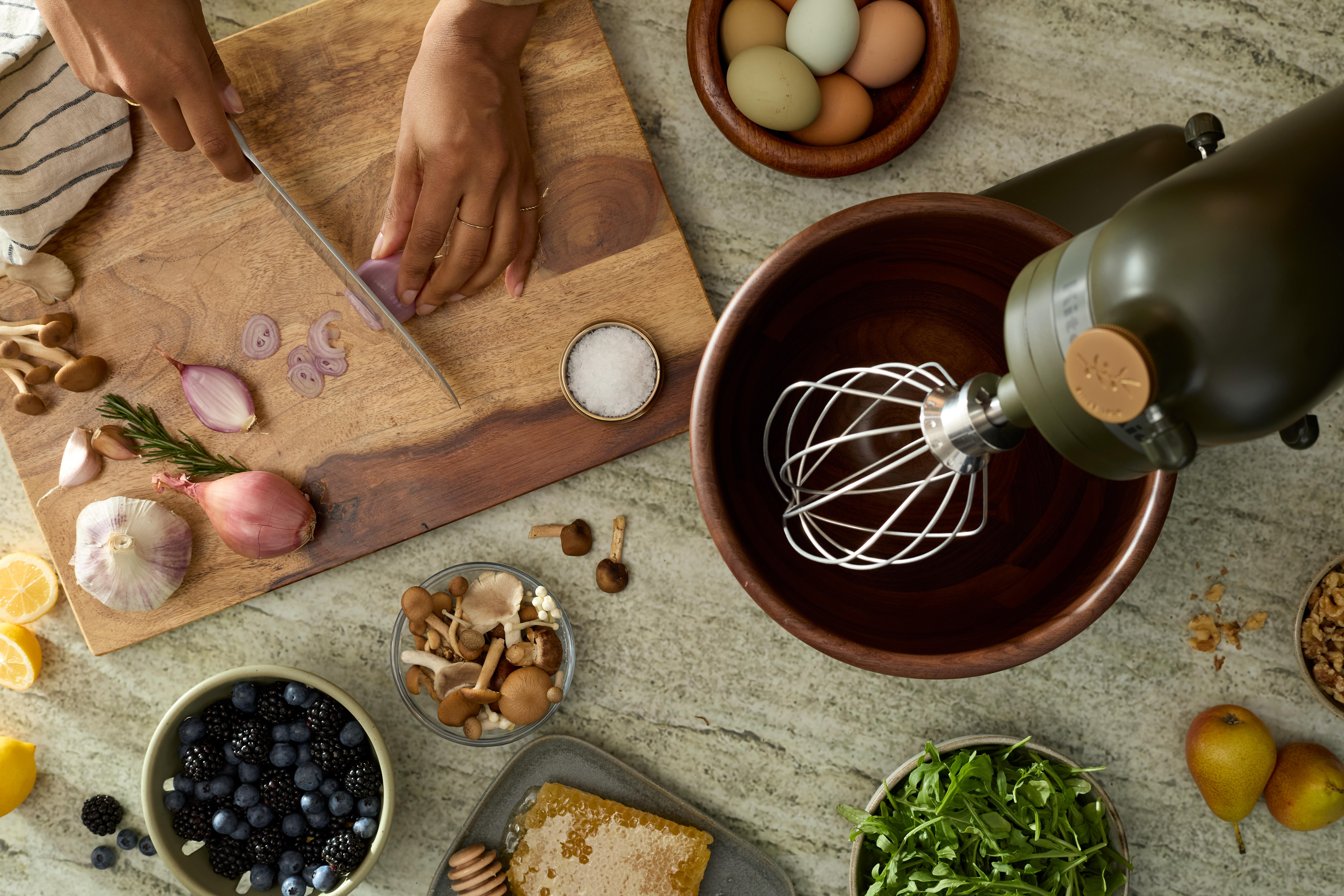 Küchenszene: Hände schneiden Schalotten, Mixer, Schüsseln mit Essen, Zutaten auf Tisch.