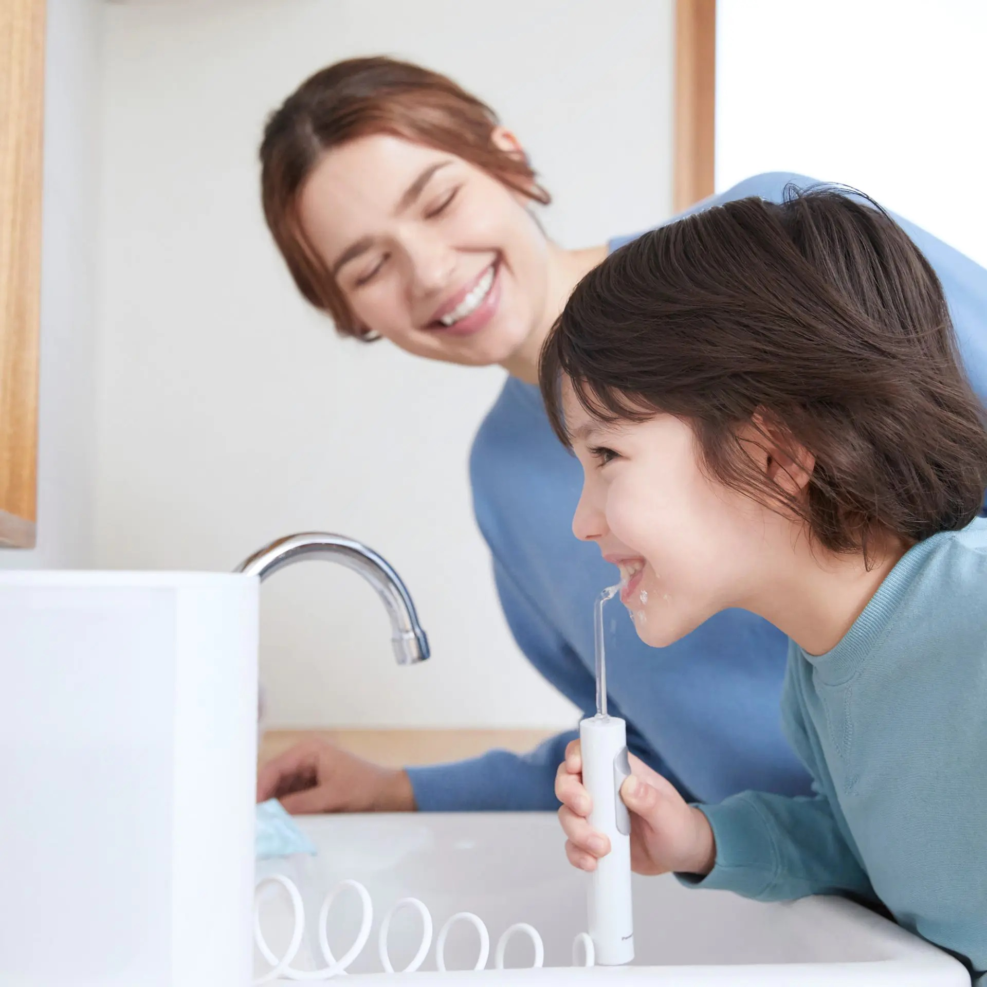 Dziecko używa irygatora do jamy ustnej, a w tle uśmiecha się kobieta w pobliżu zlewu.
