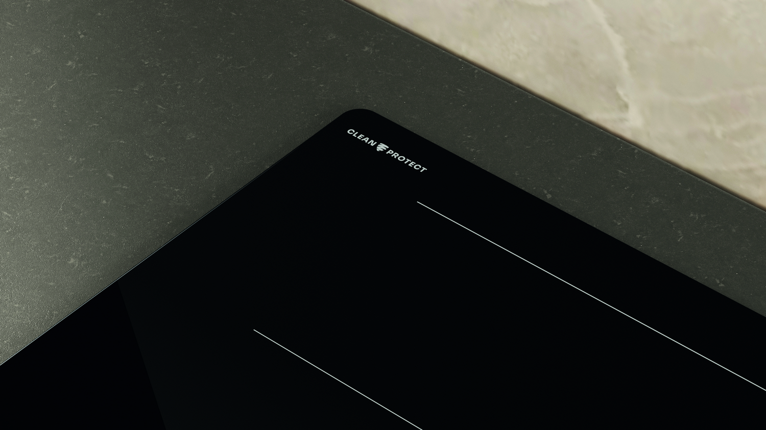 Primer plano de una placa de inducción negra con líneas blancas y texto "CLEAN PROTECT". Superficies gris oscuro y beige en el fondo.