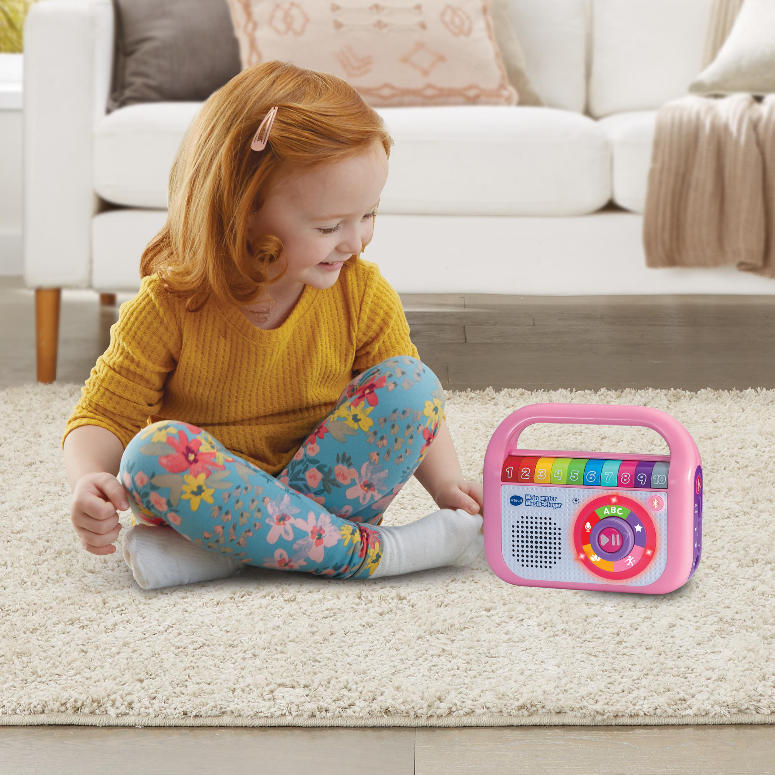 Ein junges Mädchen sitzt auf einem Teppich und spielt mit einem rosa Spielzeugradio. Sie lächelt und ist glücklich.