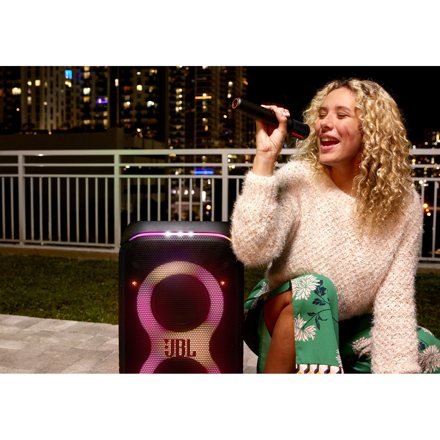 A woman singing into a microphone with a speaker, city lights in the background.