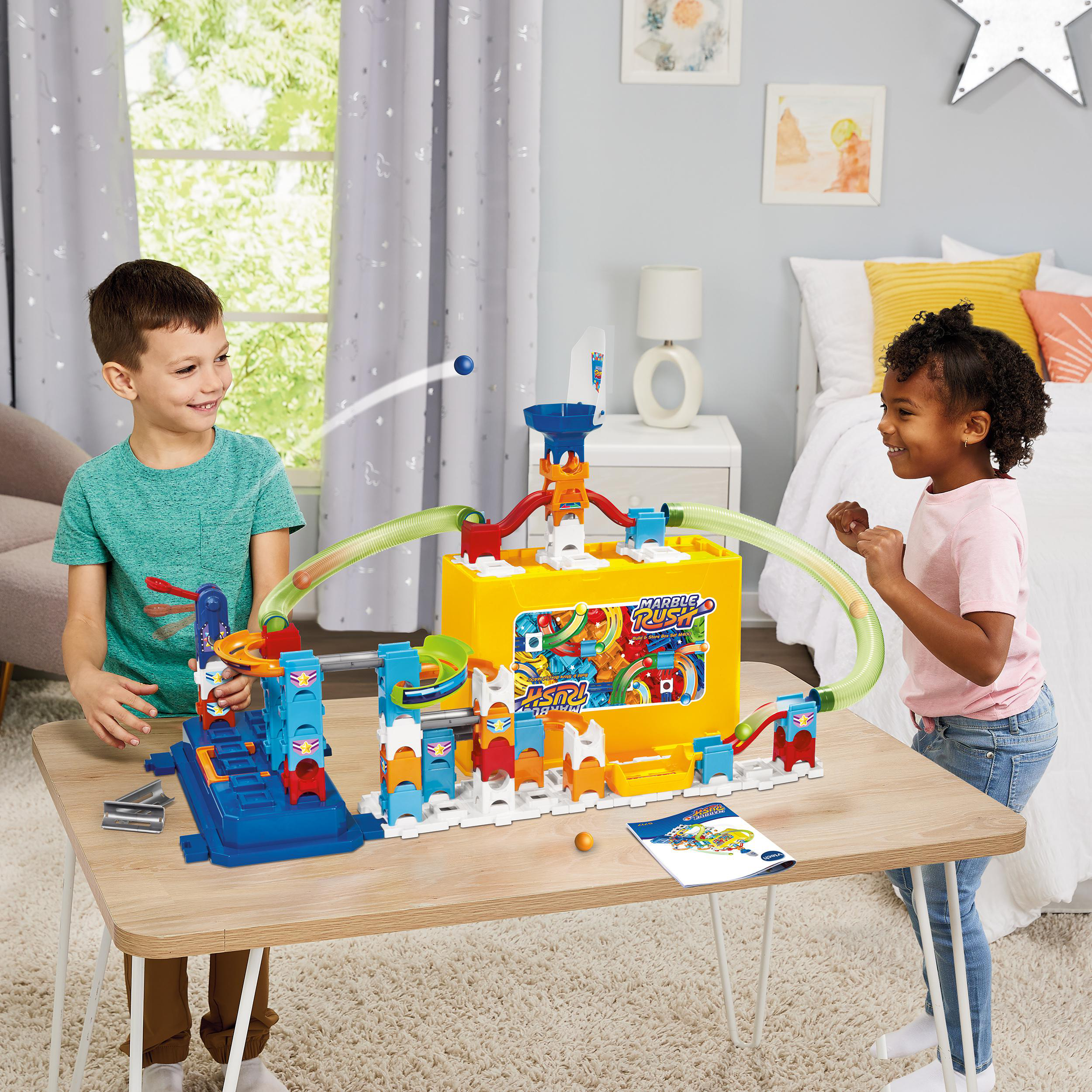 Zwei Kinder spielen mit einem Murmelbahn-Spielzeug auf einem Tisch in einem Schlafzimmer.