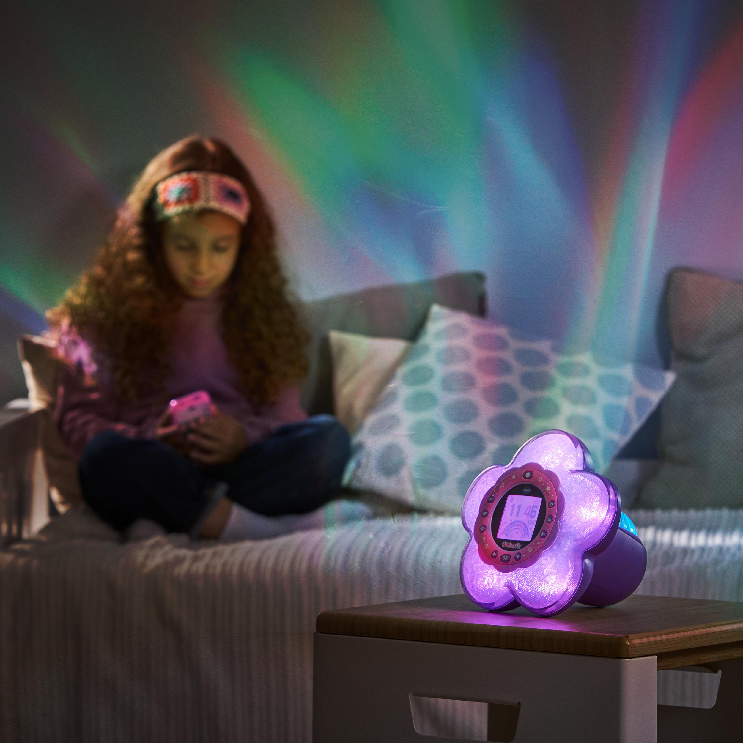 Mädchen mit Handy am Bett, beleuchtet von einem blumenförmigen, violetten Wecker. Buntes Licht an der Wand.