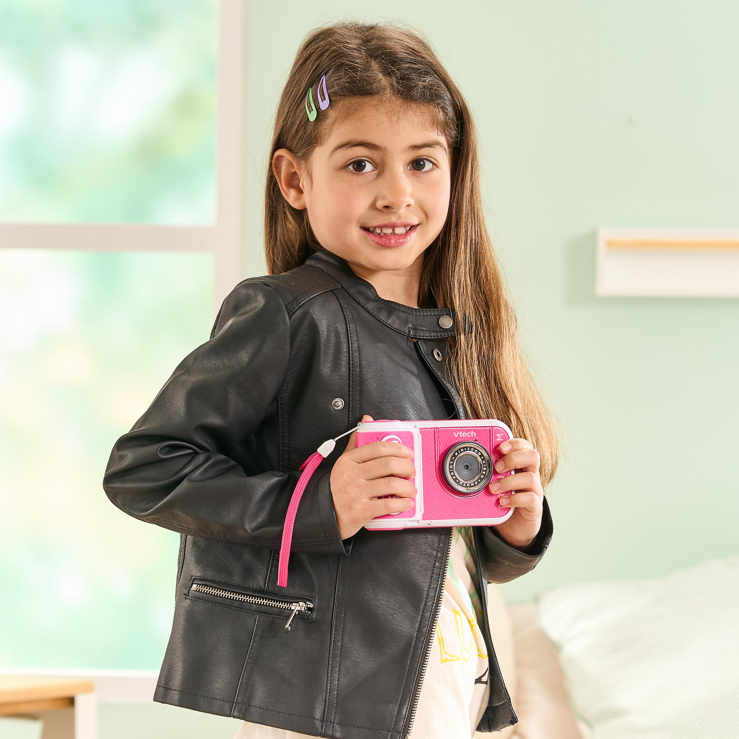 Ein Mädchen hält eine rosa Spielzeugkamera und trägt eine schwarze Jacke. Sie lächelt.