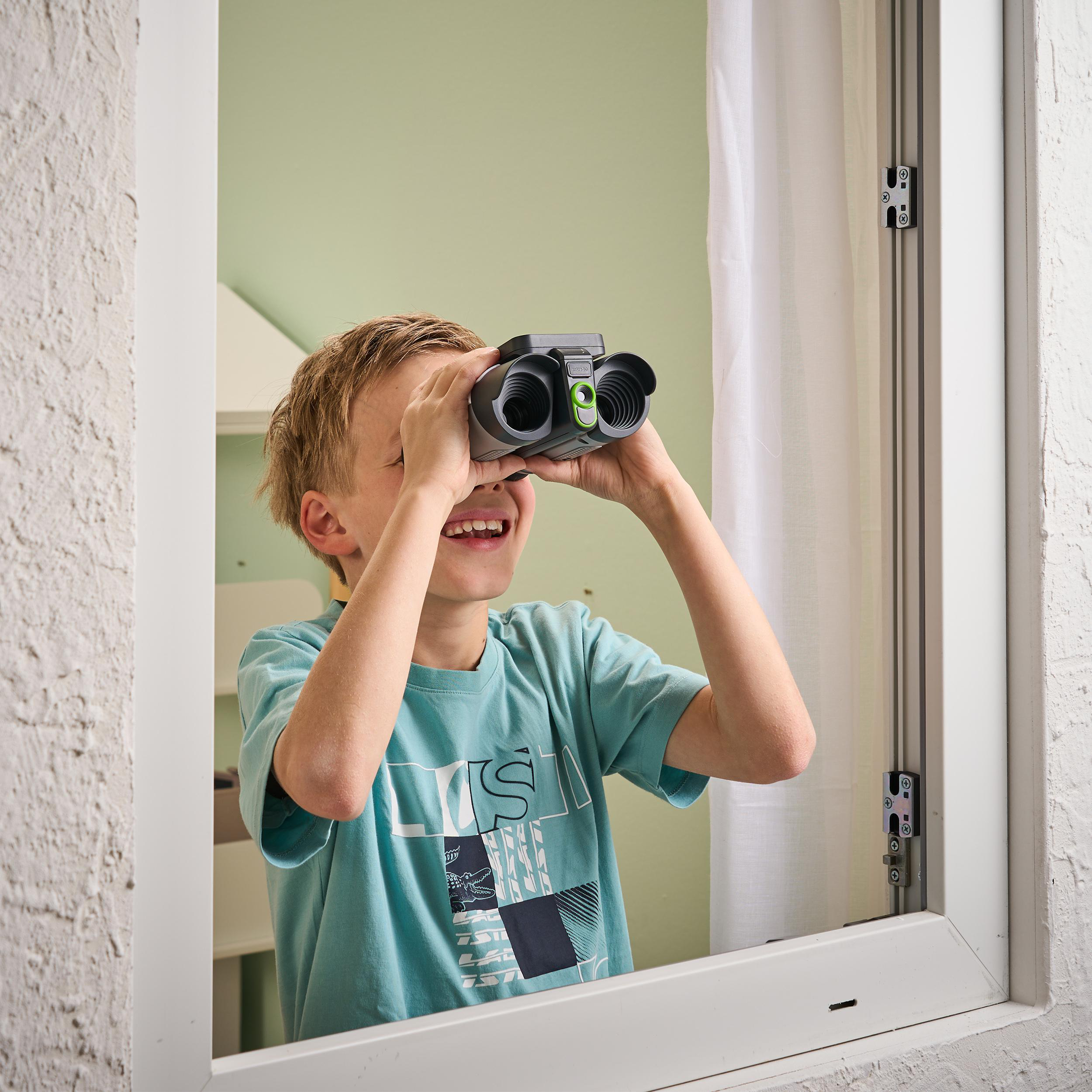 Ein Junge schaut durch ein Fernglas im Fenster und lächelt. Grünes Hemd, hellgrüne Wand.
