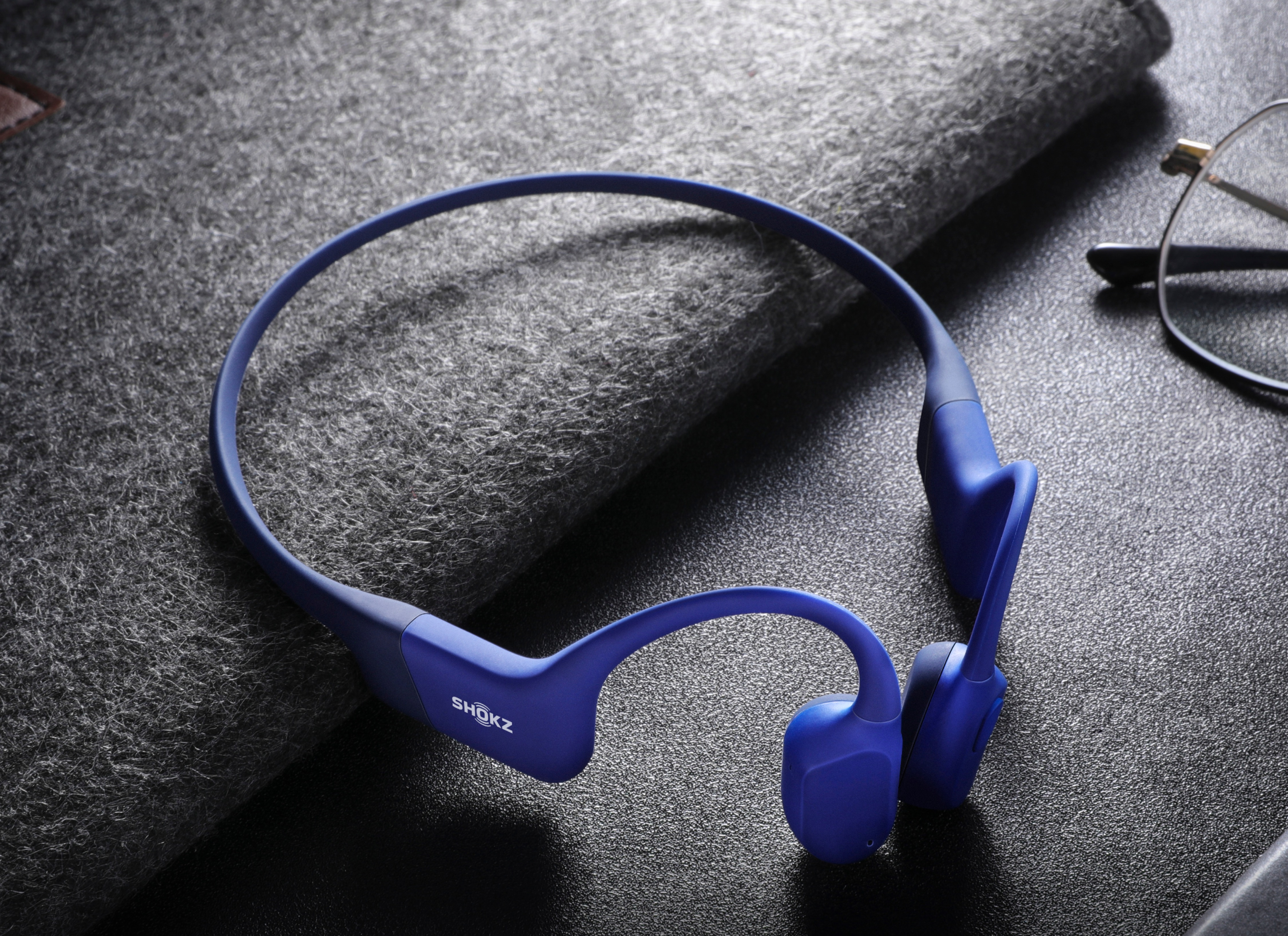 Casque Shokz bleu sur une surface grise, avec des lunettes en arrière-plan.