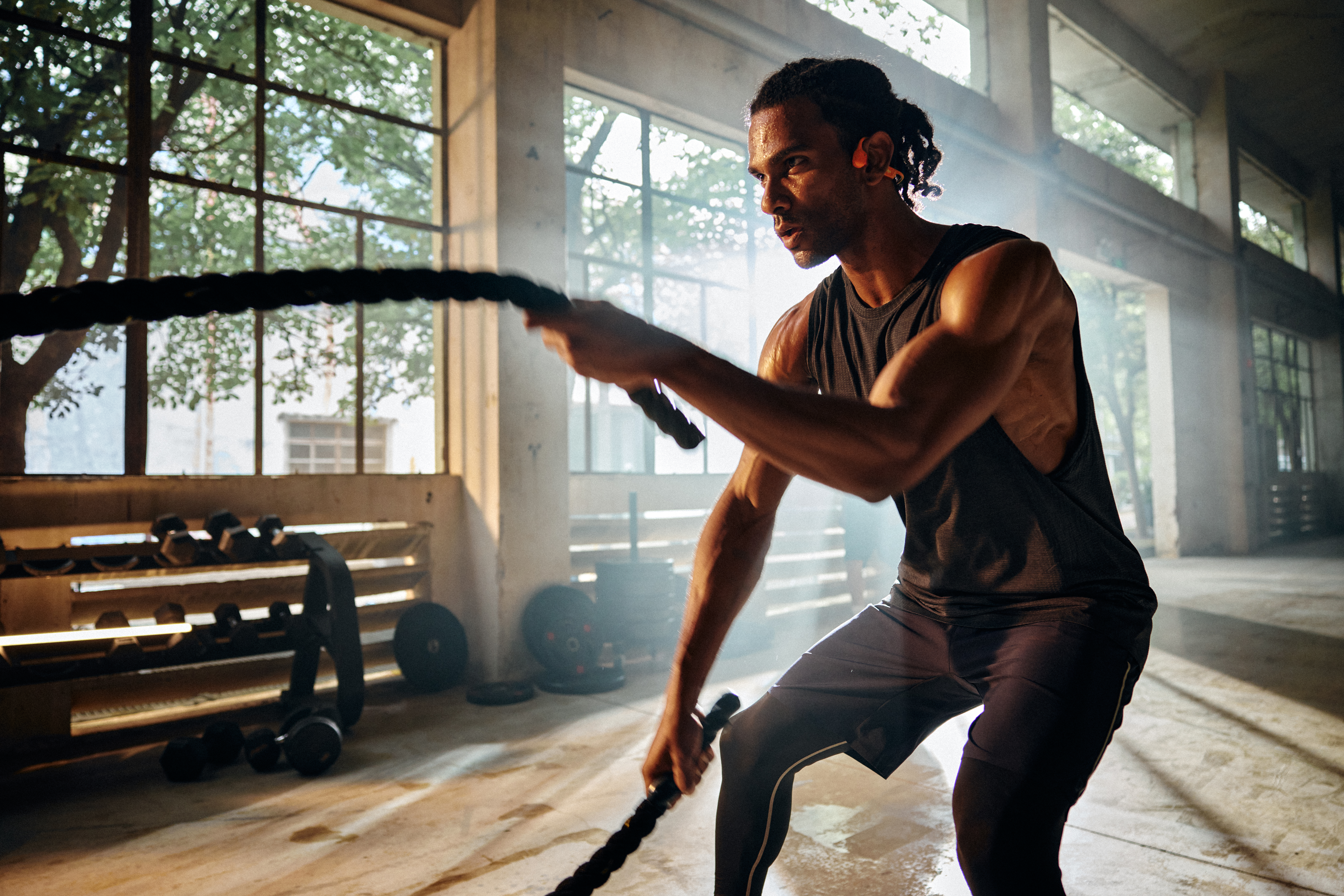 Ein Mann zieht im Fitnessstudio ein Battle Rope. Sonnenlicht scheint durch Fenster und beleuchtet die Szene. Hanteln im Hintergrund.