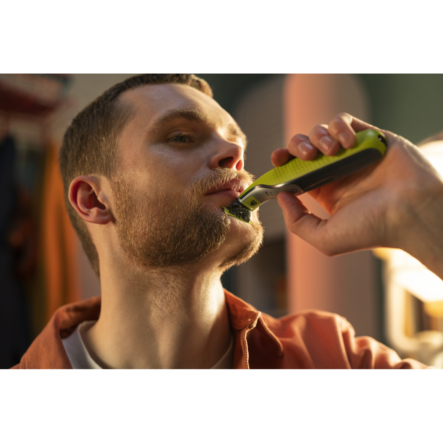 A man is shaving his beard with a green and black electric shaver, in a close-up shot.