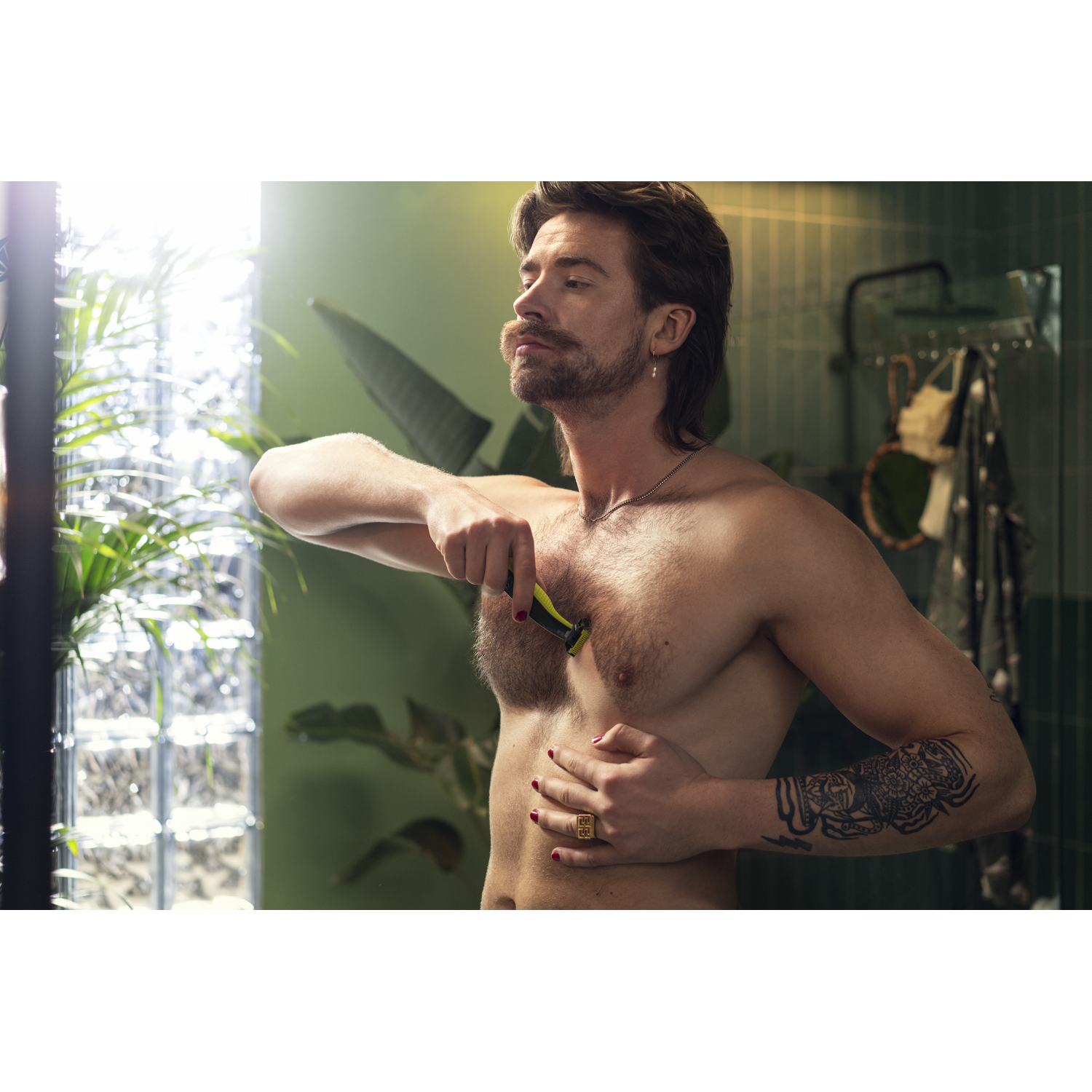 A man shaves his chest with a yellow trimmer, with tattoos and red nail polish.