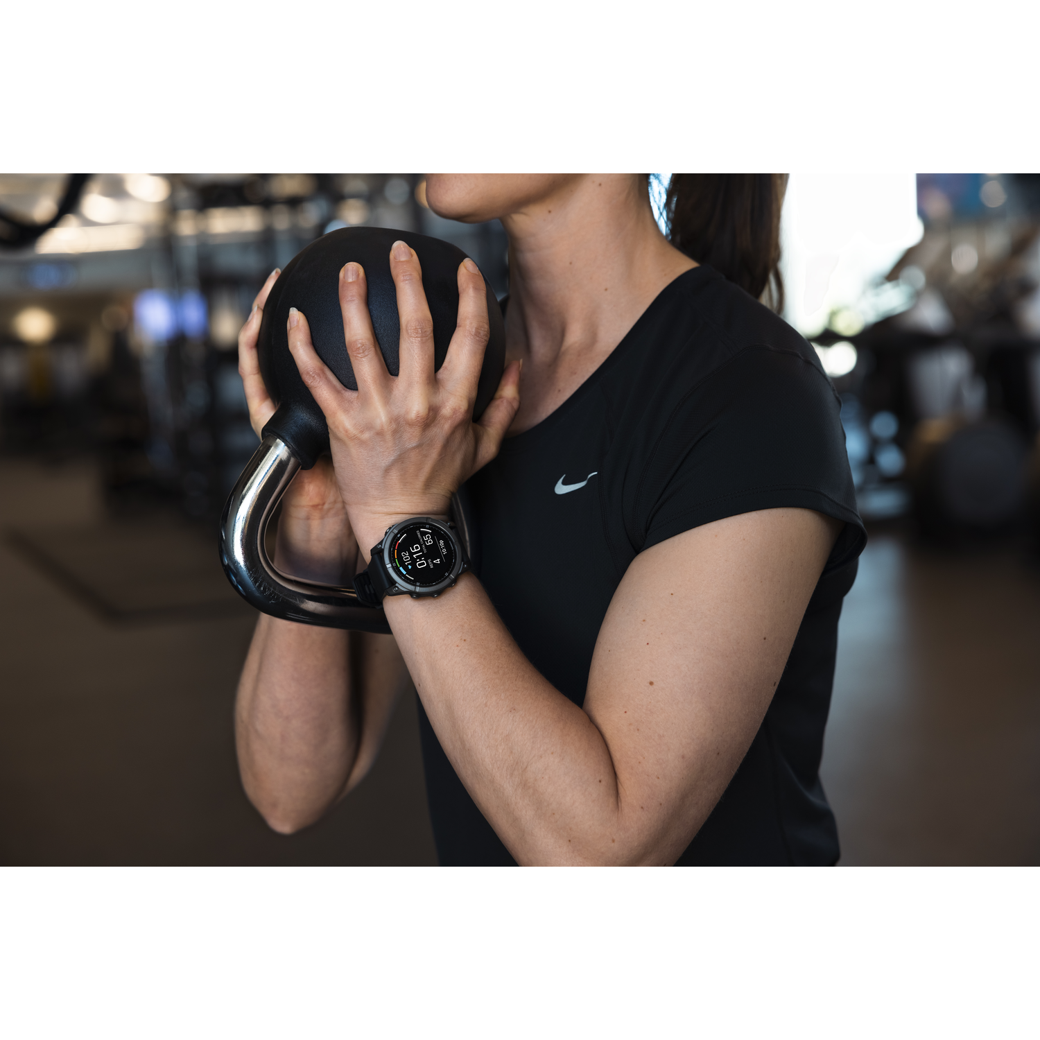 A woman is holding a kettlebell and wearing a smartwatch. She is in a gym.
