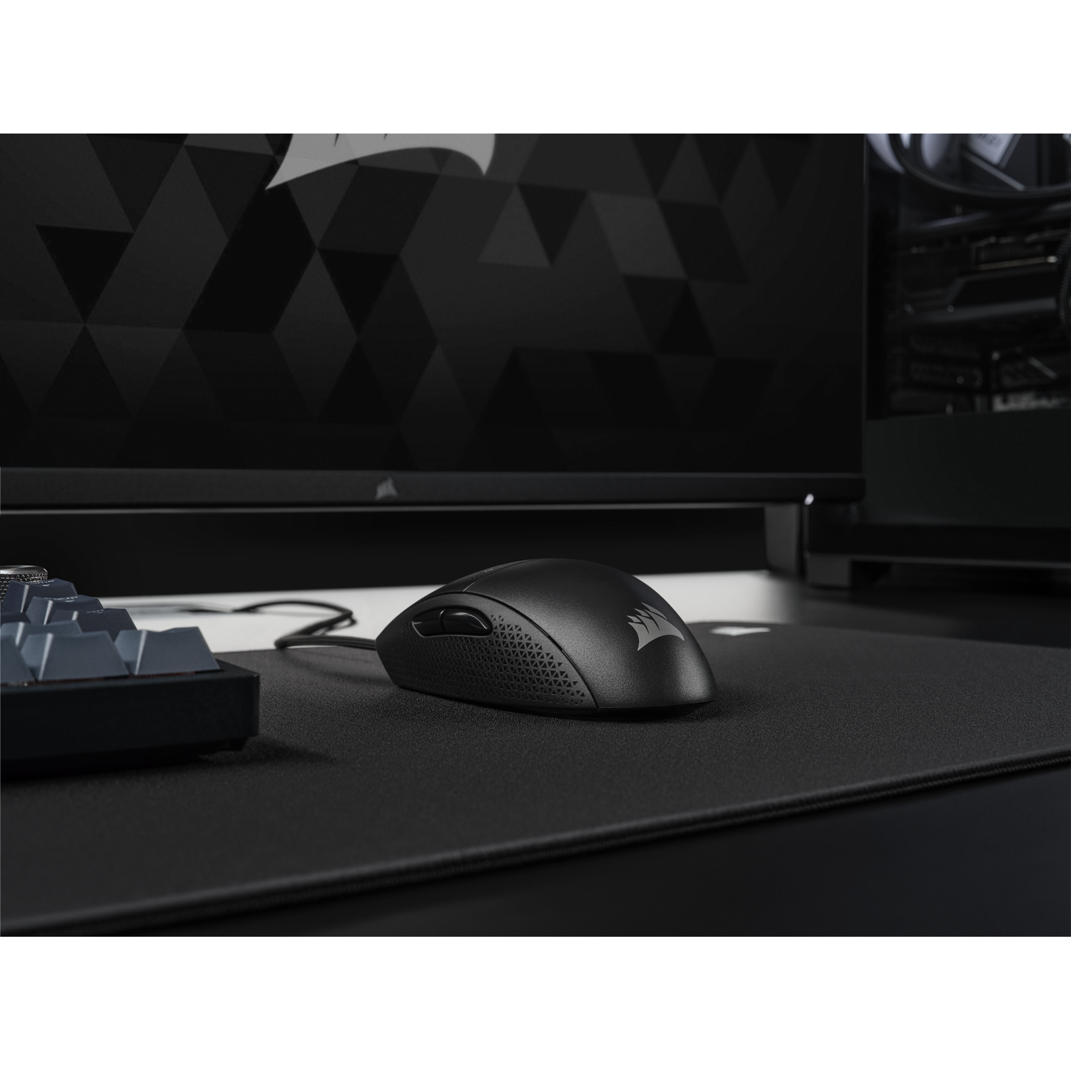 Black gaming mouse on a black mousepad, with keyboard and monitor in the background.
