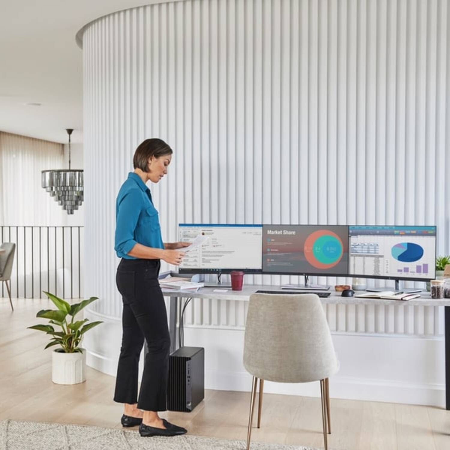 Vrouw in blauw overhemd staat, kijkt naar computerschermen op een bureau.