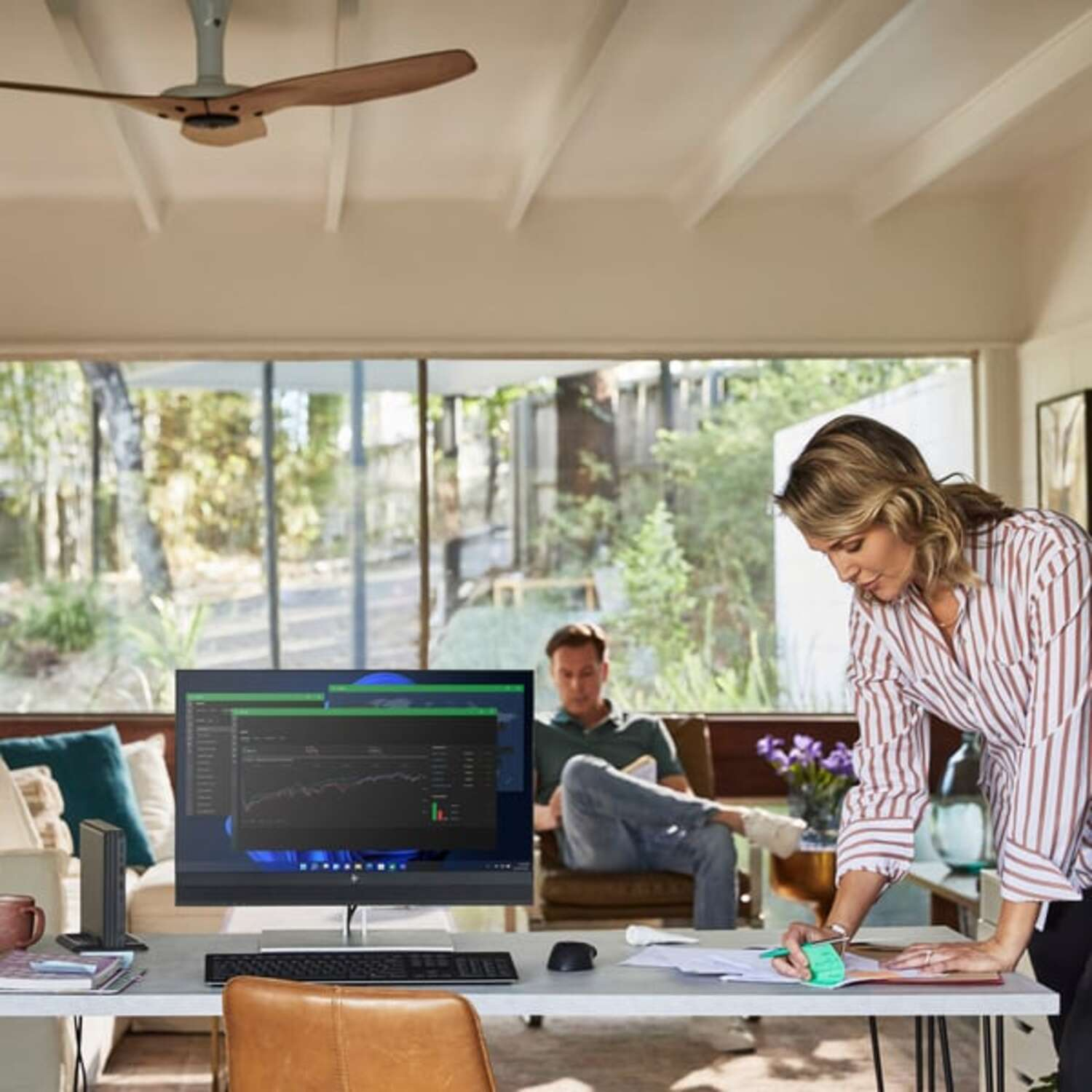 Vrouw aan bureau, man op bank, computerscherm met grafieken, modern interieur.