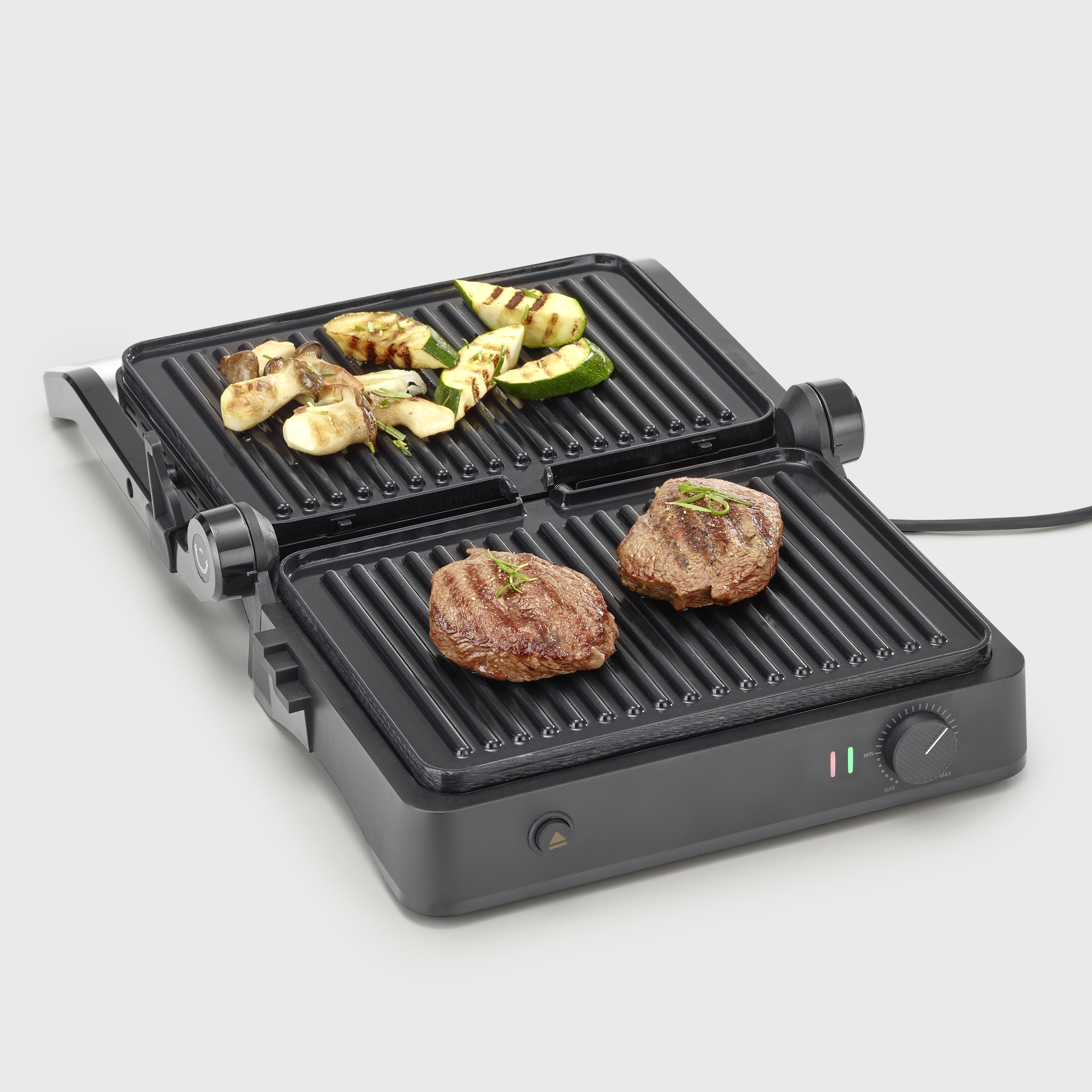 Offener Grill mit gegrilltem Steak und Gemüse. Schwarzer Grill auf weißer Oberfläche.