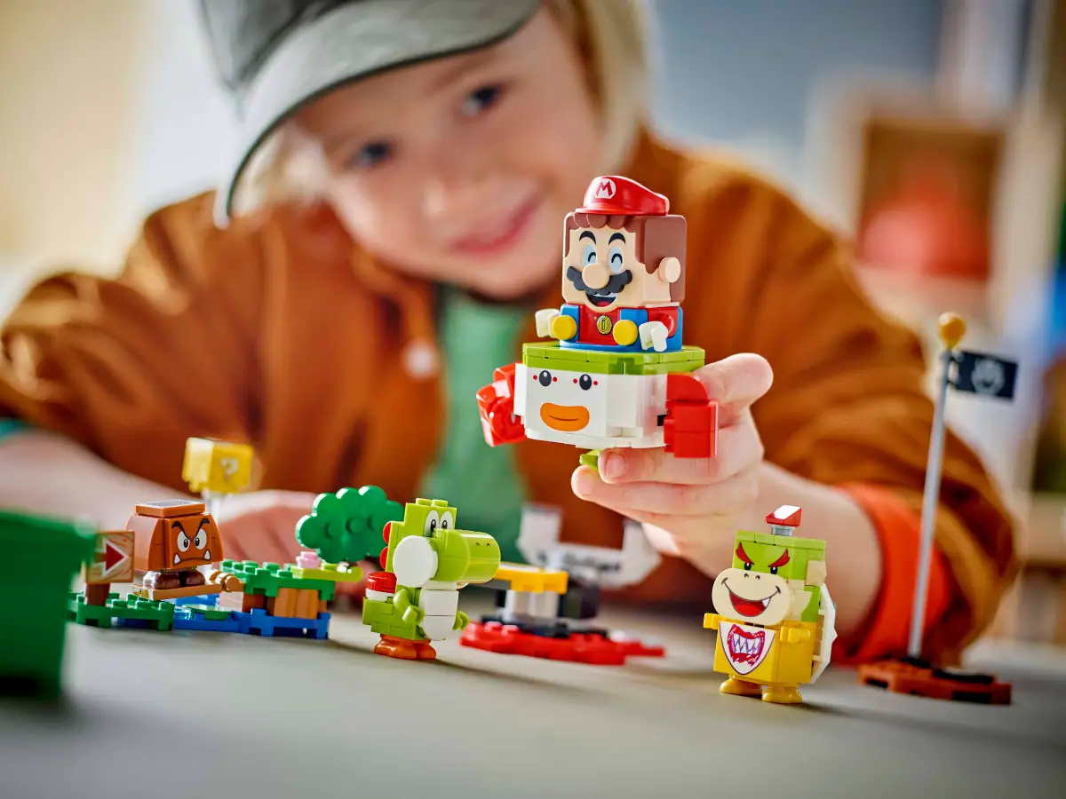 Dziecko bawiące się zestawem Lego Mario, w tym figurka Mario, Yoshi i Bowser Jr. na szarej powierzchni.