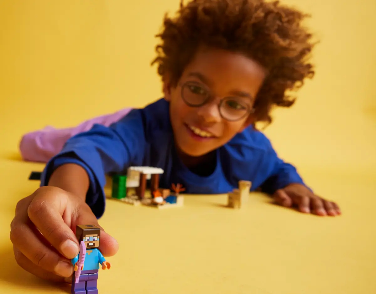 Chłopiec w okularach bawiący się figurką LEGO Minecraft, żółte tło.