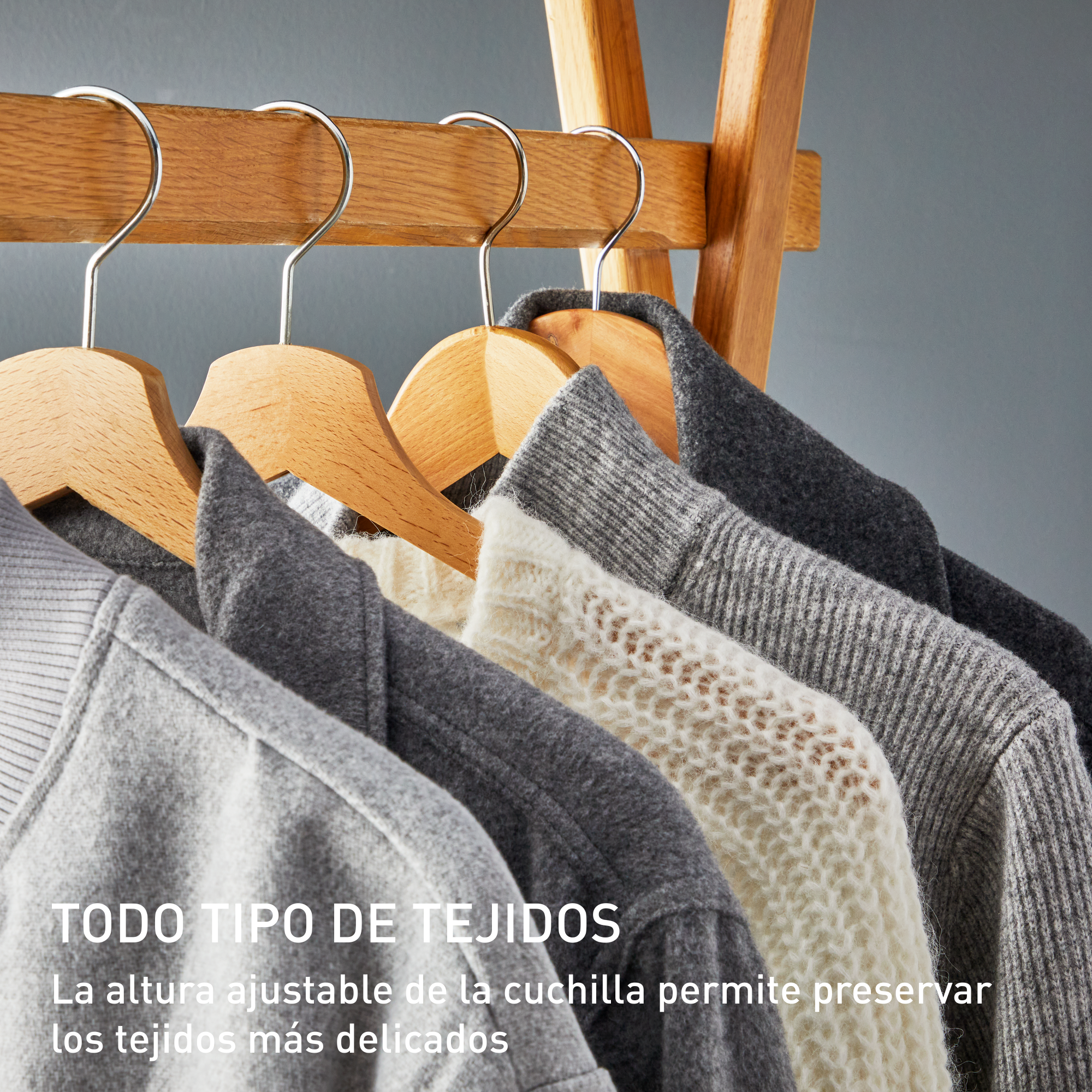 Ropa colgada en perchas de madera. Varios suéteres grises y blancos. Estante de madera y fondo gris.
