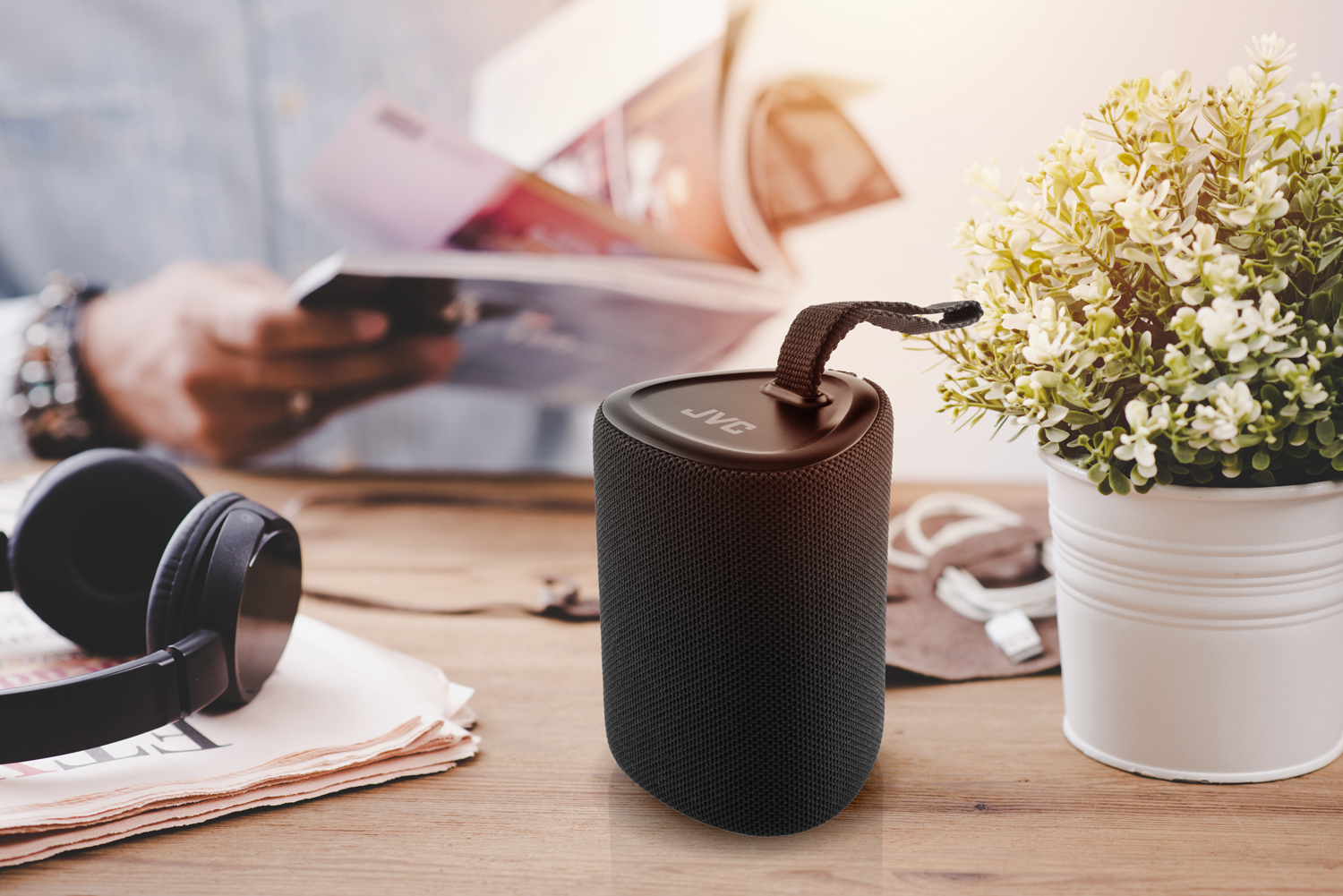 Czarny głośnik przenośny, roślina i słuchawki na drewnianym stole. Osoba czyta czasopismo.