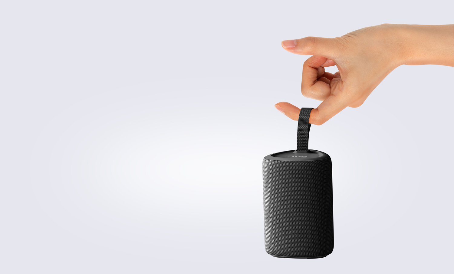 Ręka trzyma czarny, cylindryczny głośnik JVC. Białe tło.