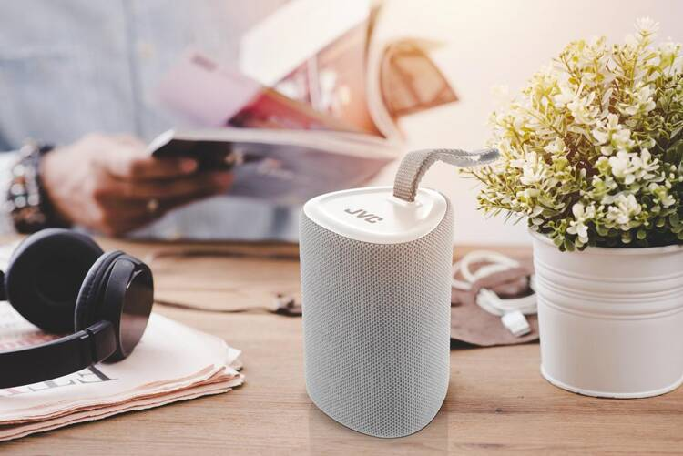 Szary głośnik JVC na drewnianym stole, z rośliną, czasopismami i słuchawkami.