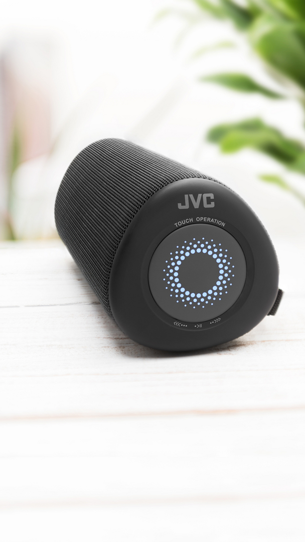 Czarny głośnik JVC na białej drewnianej powierzchni. Głośnik ma elementy sterowania dotykowego.