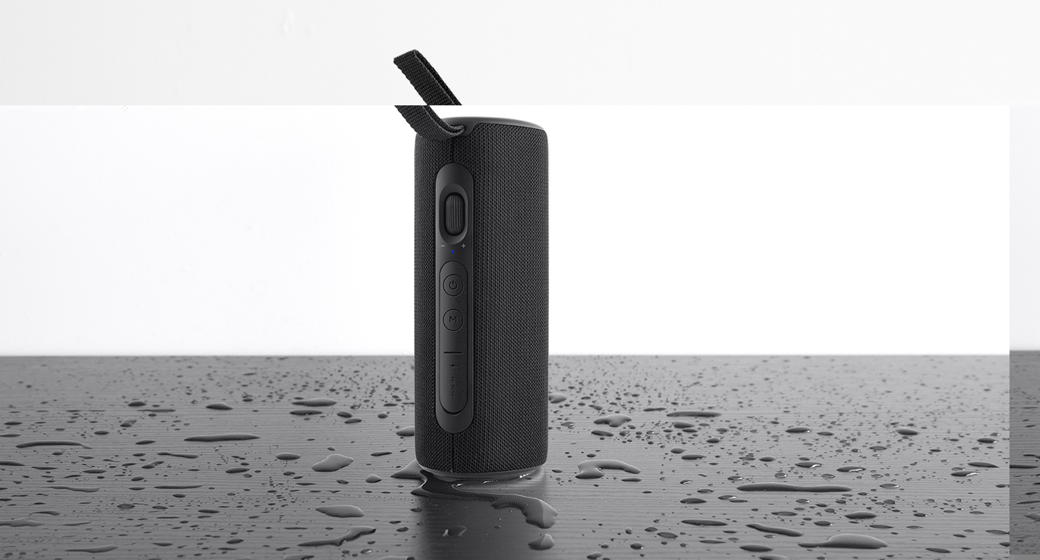 Czarny głośnik na mokrej powierzchni z kroplami wody. Białe tło, widoczne przyciski boczne.