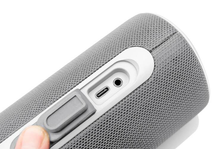Widok z boku szarego głośnika. Porty i przycisk z boku. Białe tło.