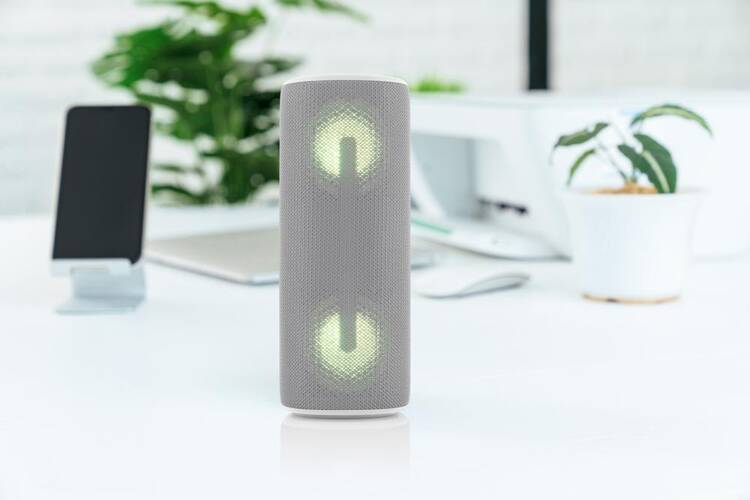 Szary głośnik na białym biurku, dwie świecące ikony zasilania. Telefon i roślina w tle.