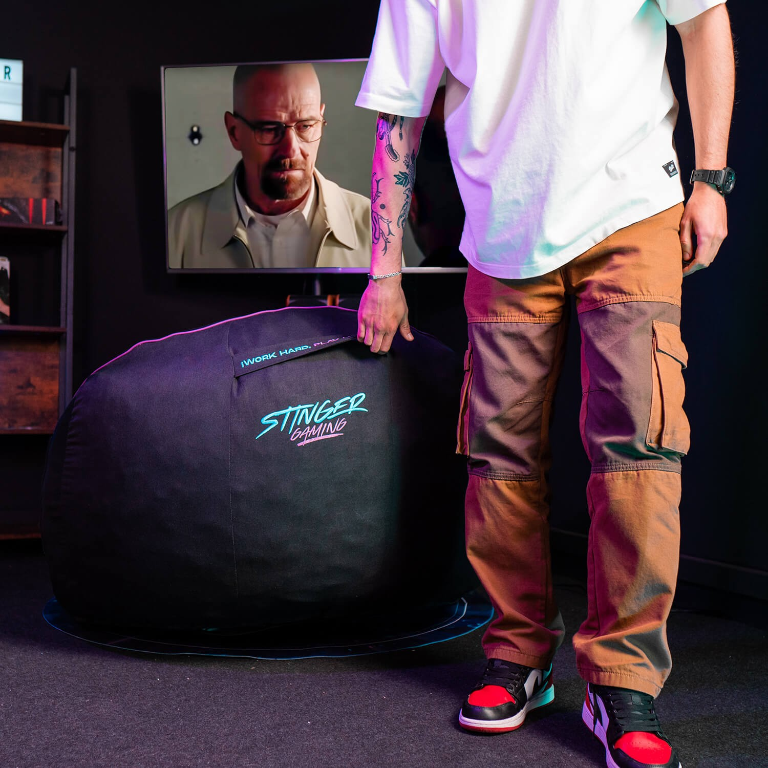 Una persona sosteniendo una silla puff Stinger Gaming, con una escena de Breaking Bad en el fondo en la TV.