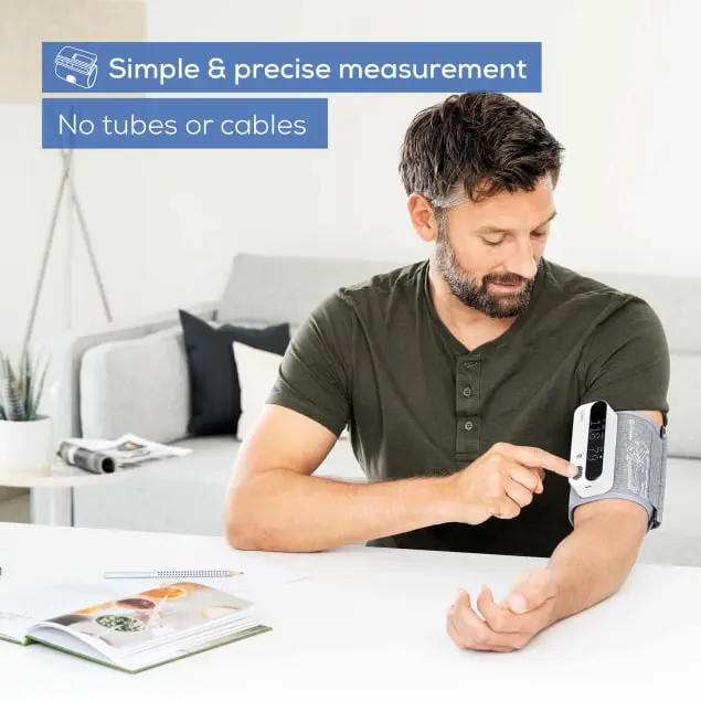 Homme utilisant un tensiomètre sur son bras. Le moniteur affiche les mesures. Table et canapé en vue.