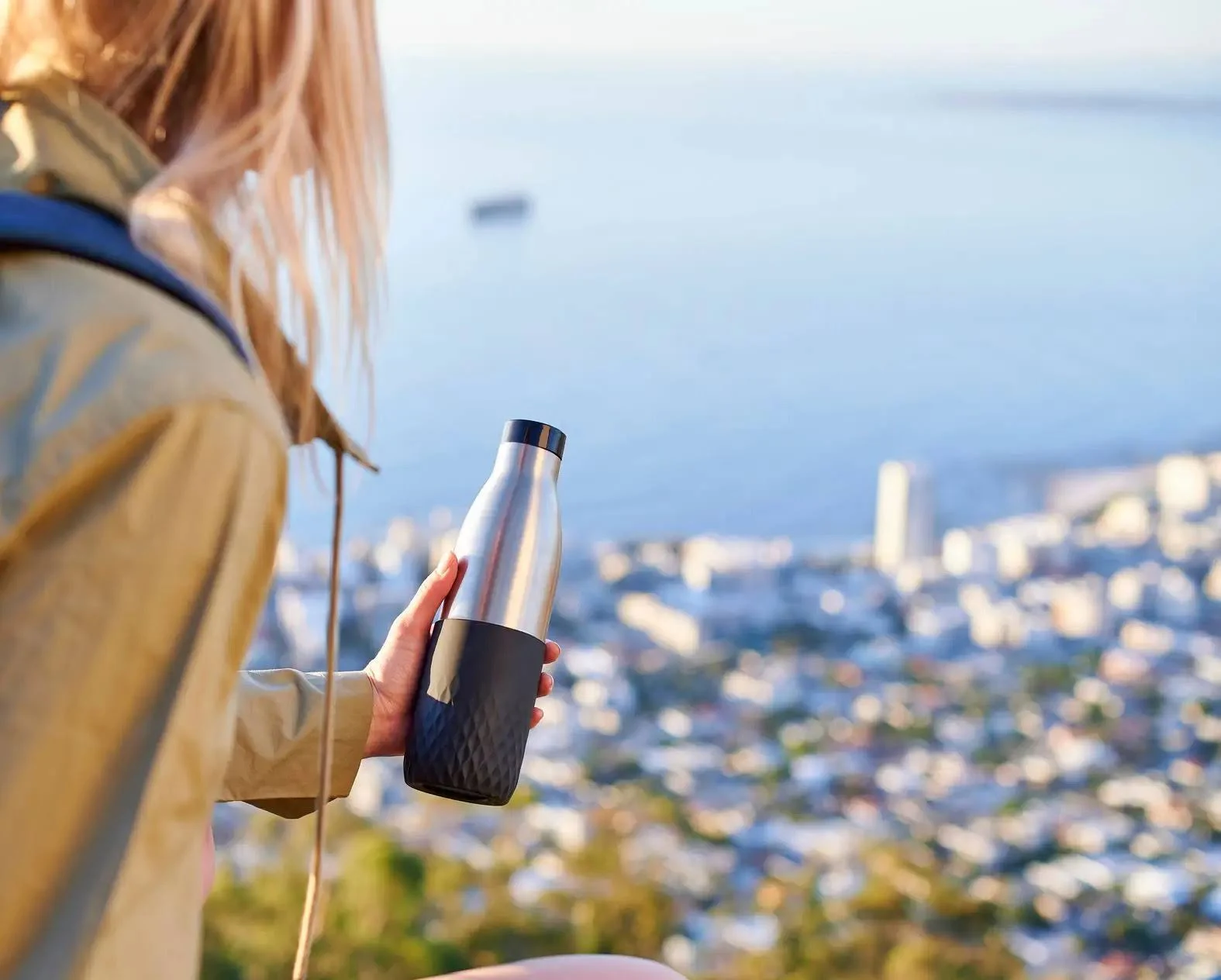 Mujer sosteniendo una botella de agua plateada y negra, con vistas a una ciudad y al mar.