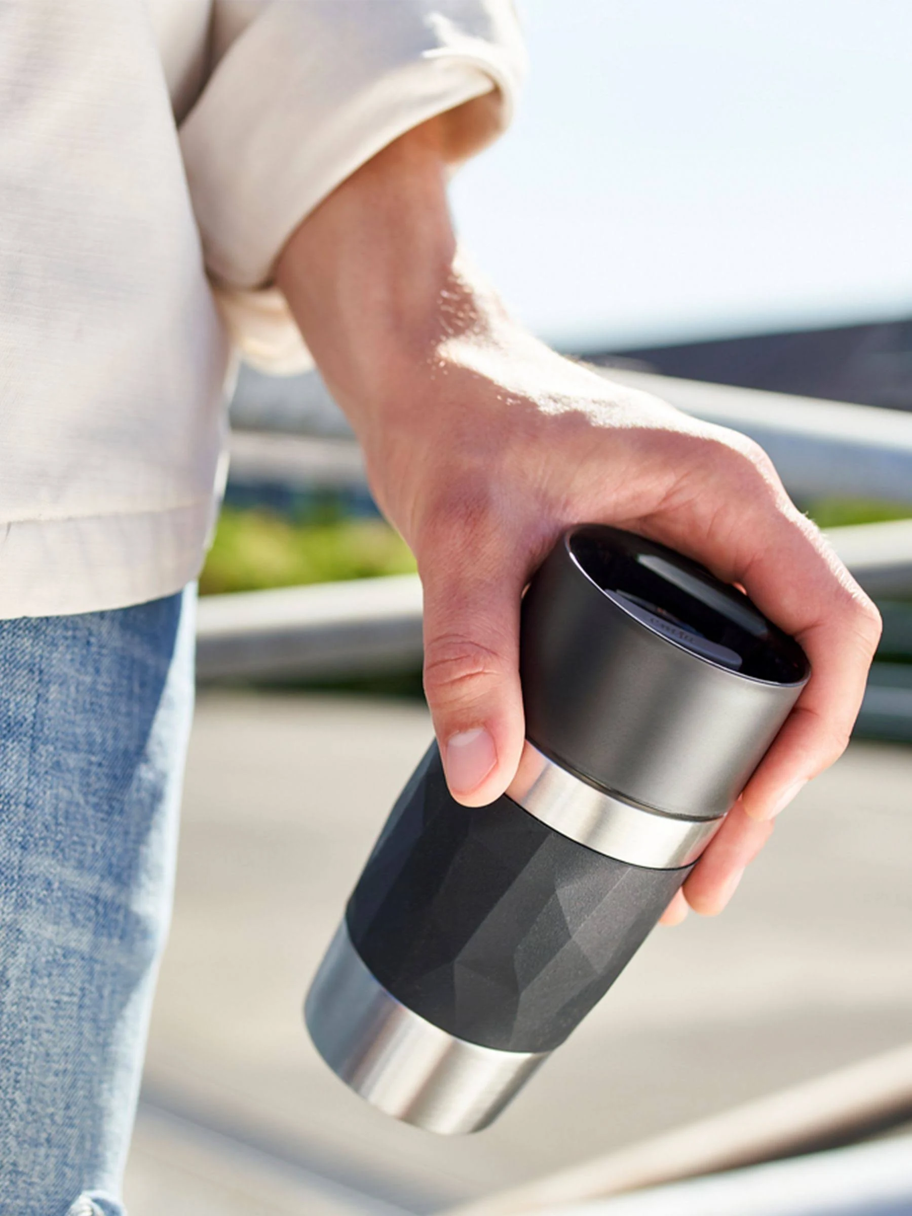 La mano de una persona sostiene un termo negro y plateado. La taza tiene un agarre negro texturizado.