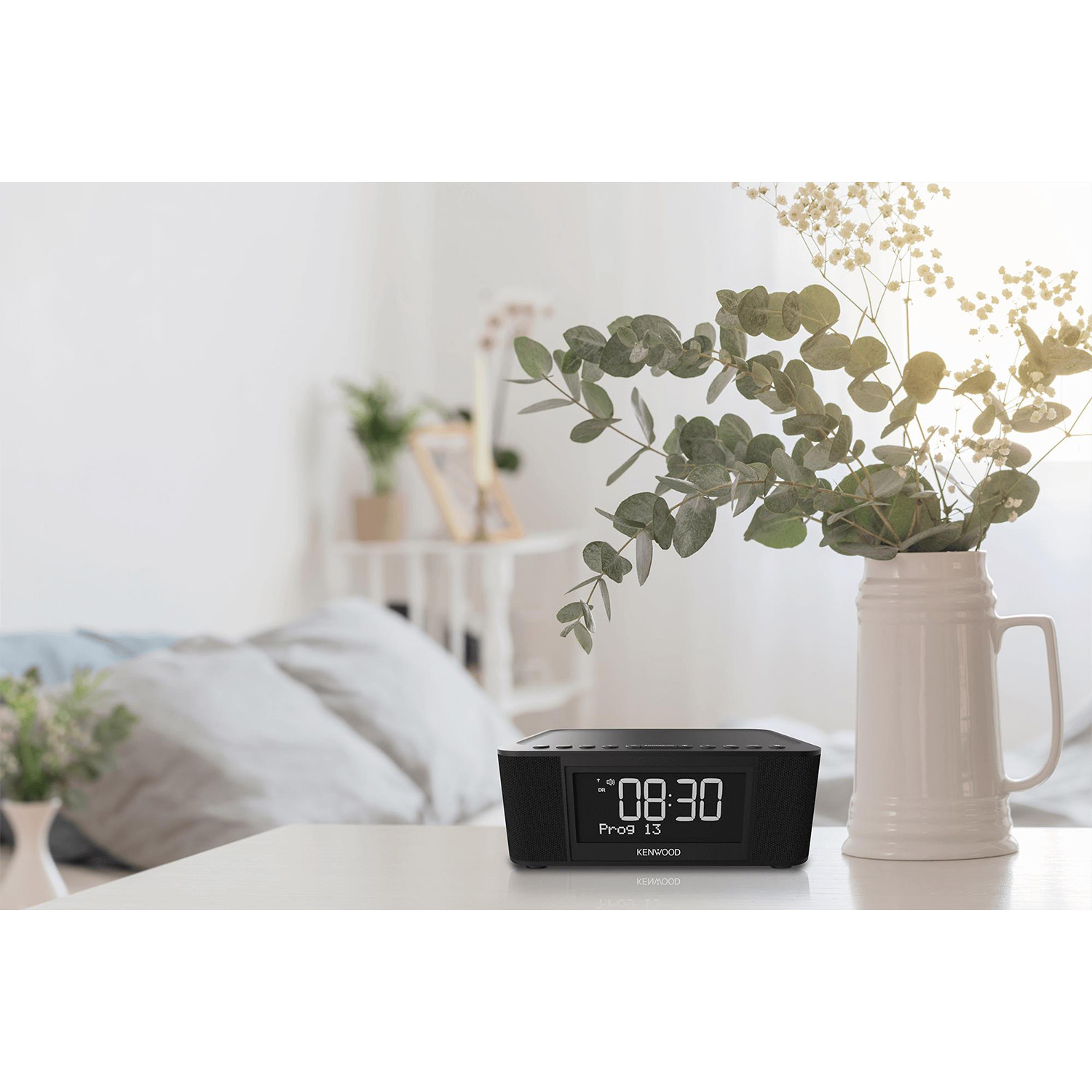 Una radiosveglia digitale nera con un vaso di fiori su un tavolo bianco. Muri bianchi.