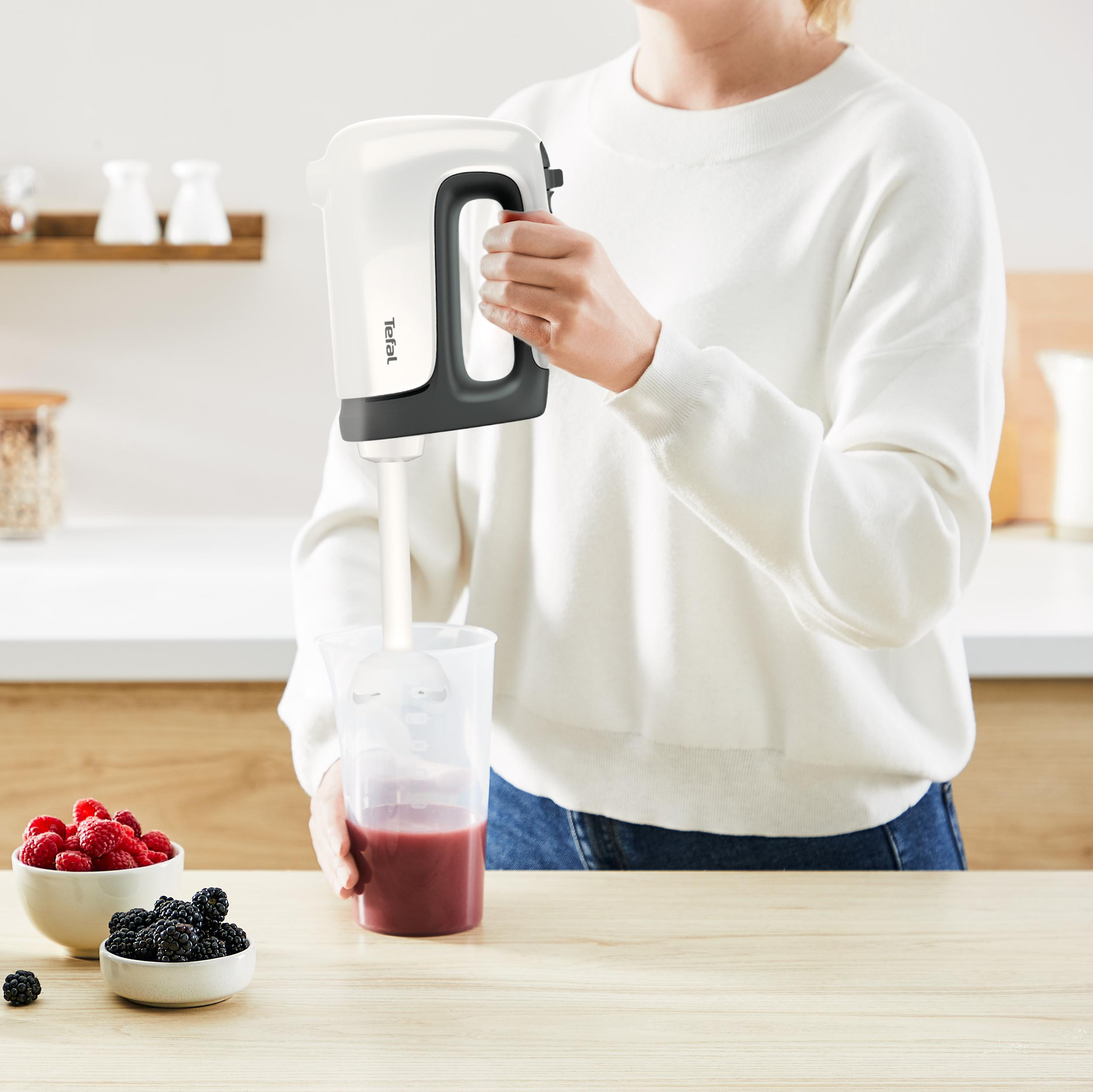 Frau, die ein rotes Getränk mit einem weißen Mixer mixt. Beeren sind in Schüsseln in der Nähe.