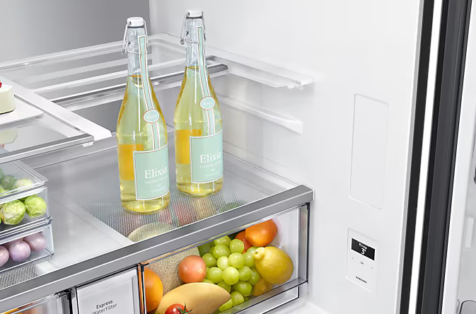 Wnętrze lodówki z dwoma butelkami, owocami i produktami. Półki są przezroczyste, drzwi otwarte.