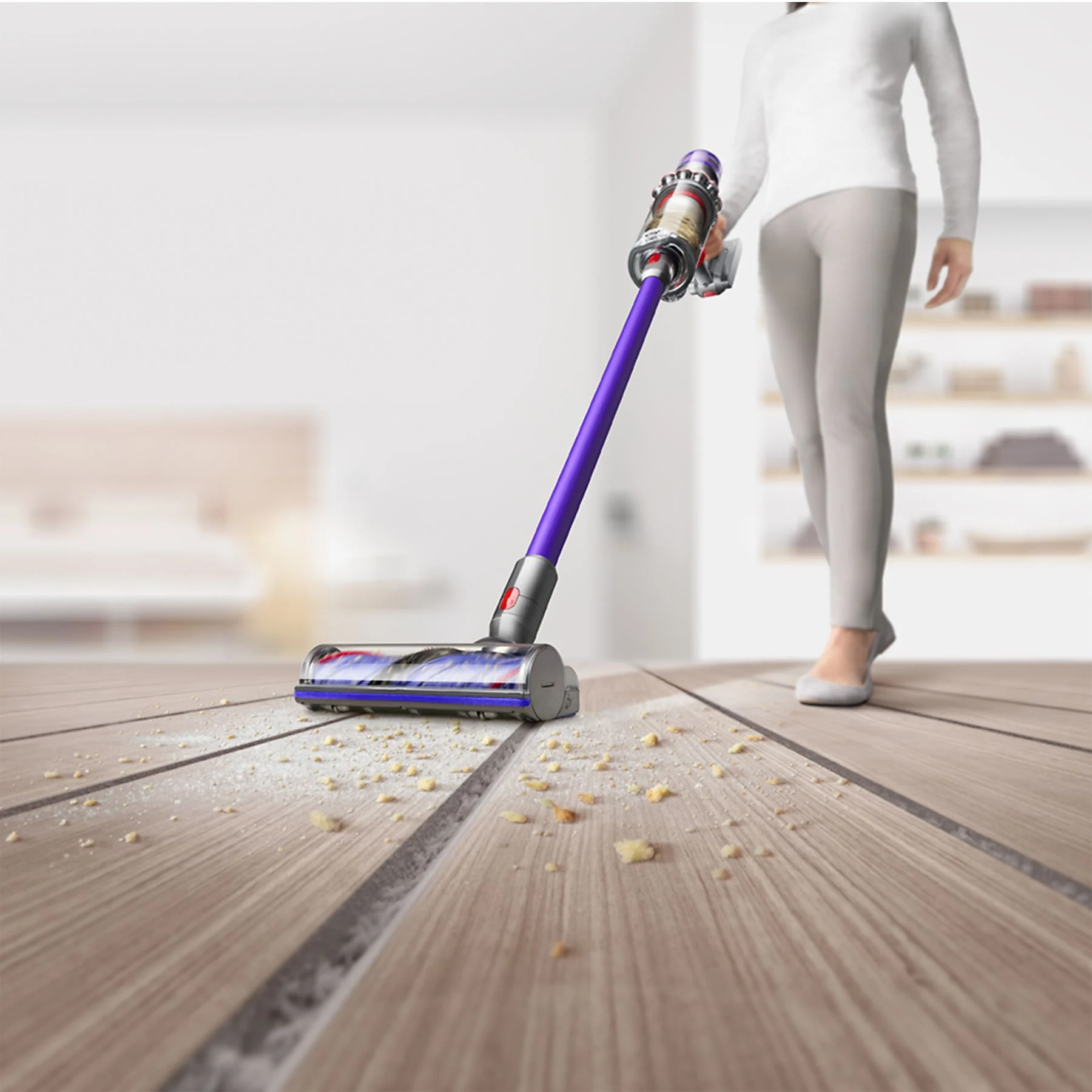 Une femme passe l'aspirateur sur un plancher en bois. L'aspirateur est un Dyson violet.