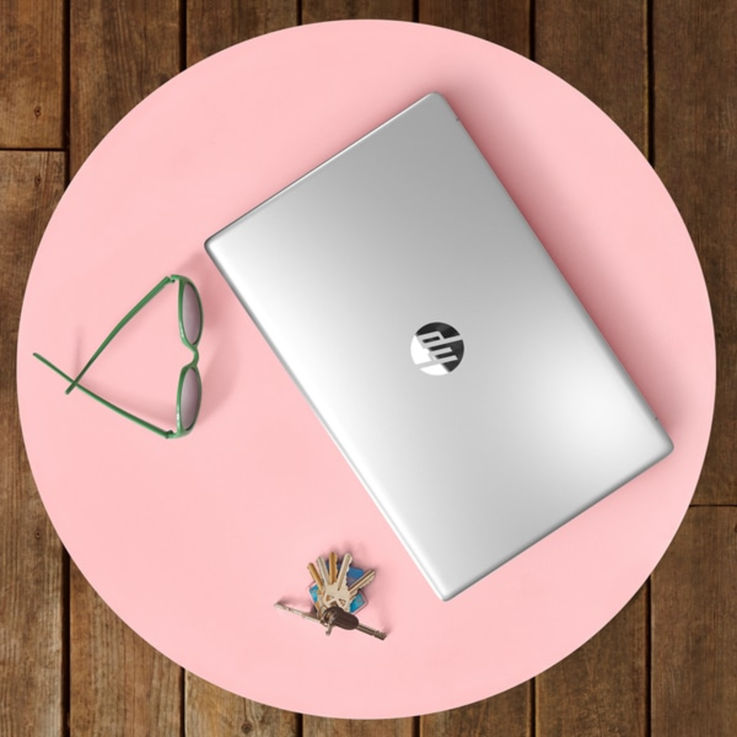 Zilveren laptop, groene bril en sleutels liggen op een roze ronde tafel, tegen een houten achtergrond.