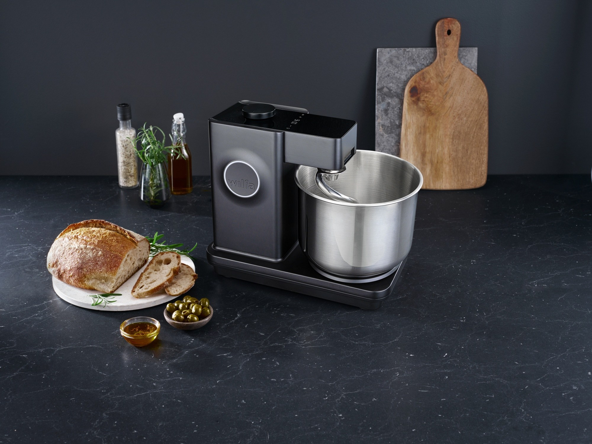 Ein schwarzer Küchenmixer mit Brot, Oliven und Öl auf dunkler Arbeitsplatte. Ein Schneidebrett ist sichtbar.