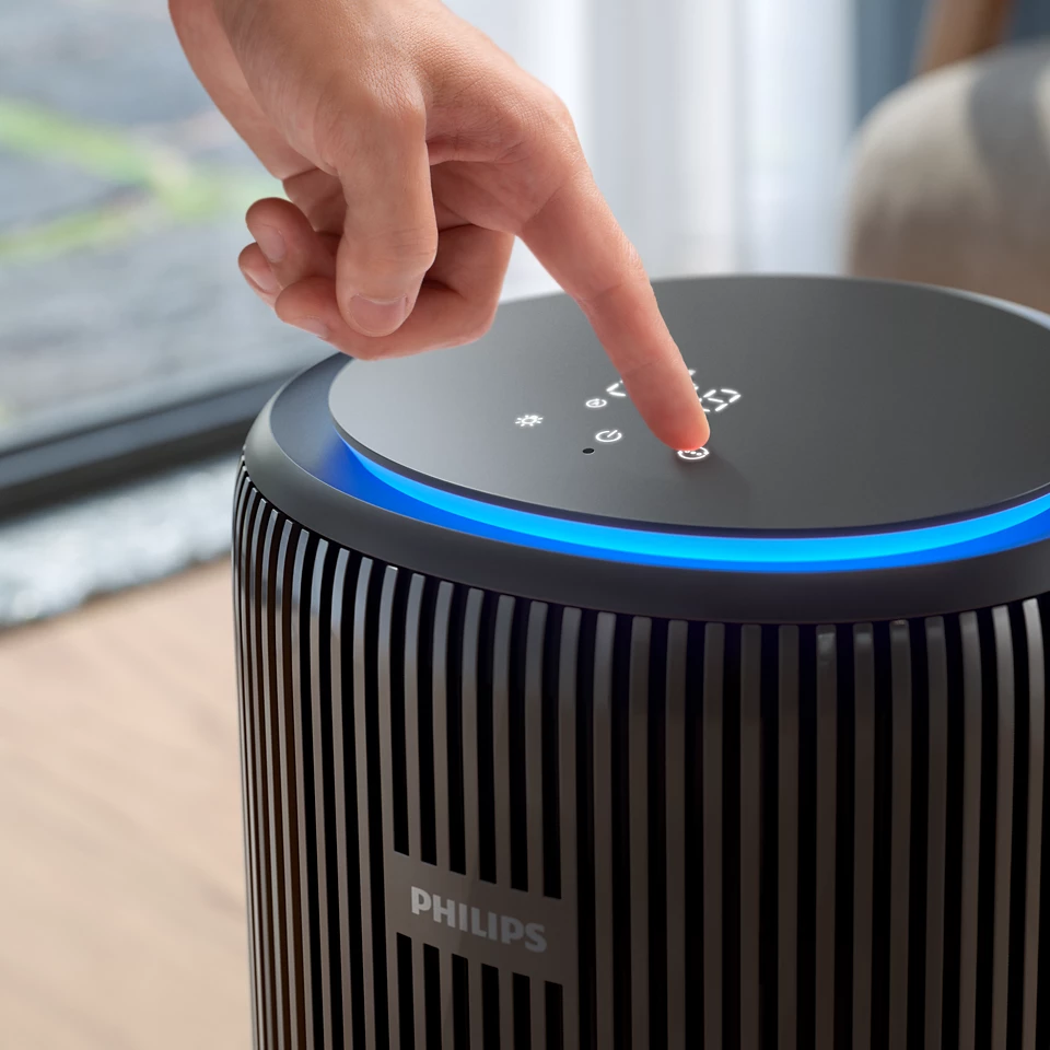 Egy személy ujja megnyom egy gombot egy fekete légtisztítón kék fénnyel. A Philips logó látható.
