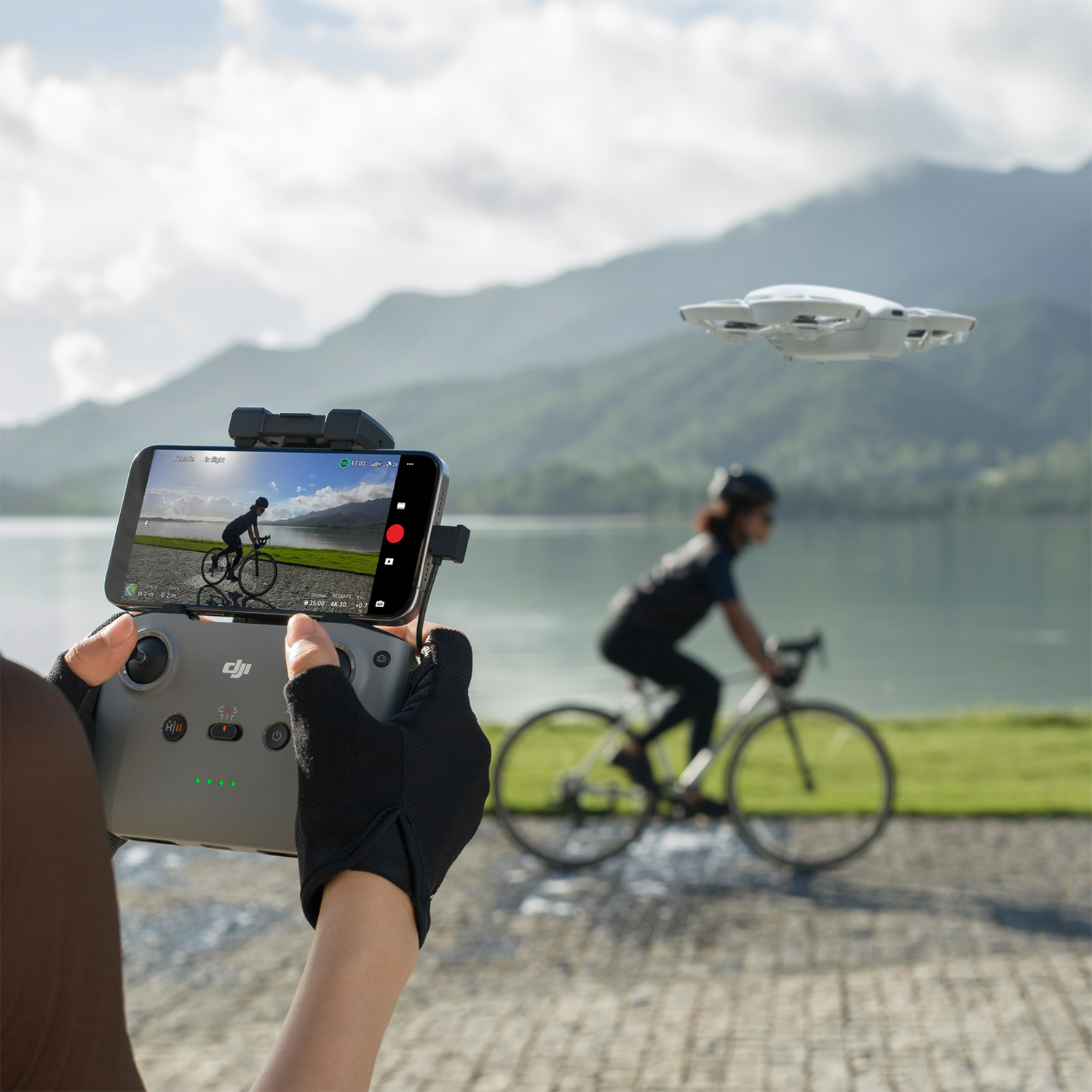 Una persona controlla un drone con uno smartphone, mentre un ciclista passa vicino a un lago.