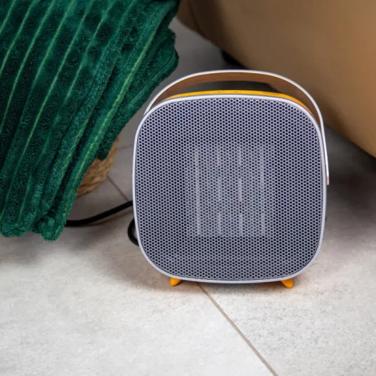 Mały, szaro-żółty grzejnik elektryczny stoi na podłodze obok zielonego koca.
