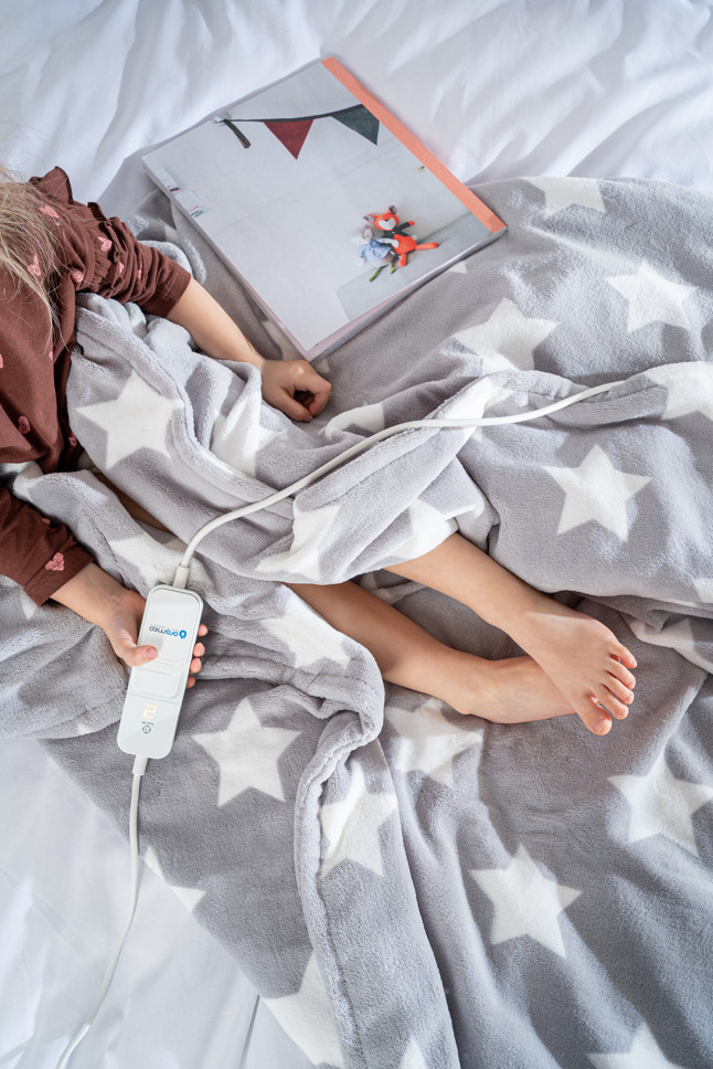 Dziecko w łóżku pod szarą kołdrą z białymi gwiazdami. Otwarta książka z zabawką na łóżku.
