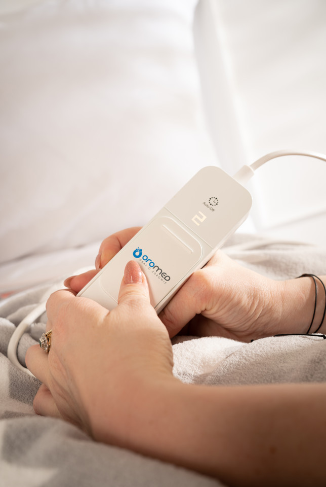 Dłonie trzymają białe urządzenie z kablem. Urządzenie ma logo i przycisk.