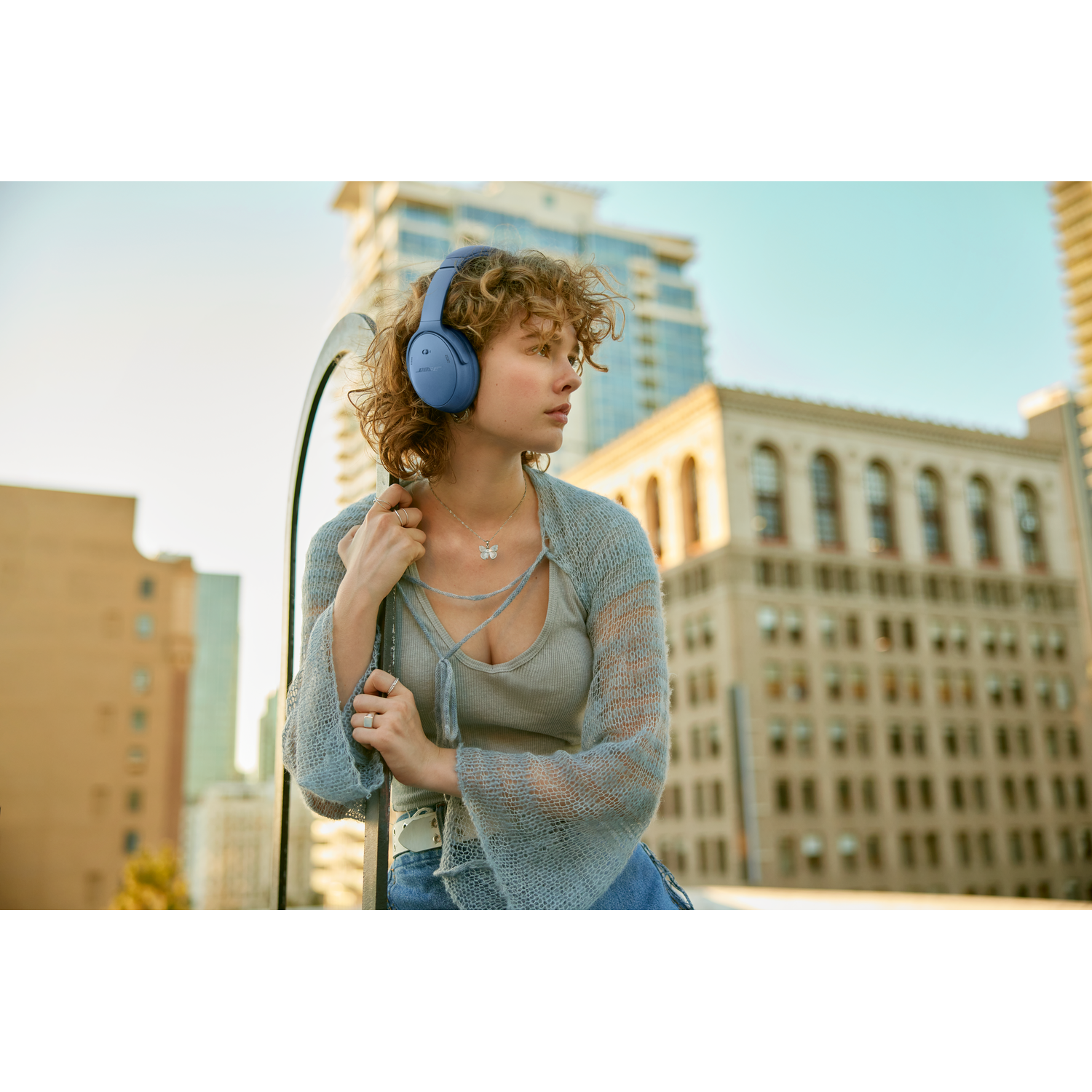 Woman with blue headphones looks away, urban background. Buildings are visible. Sunny day.