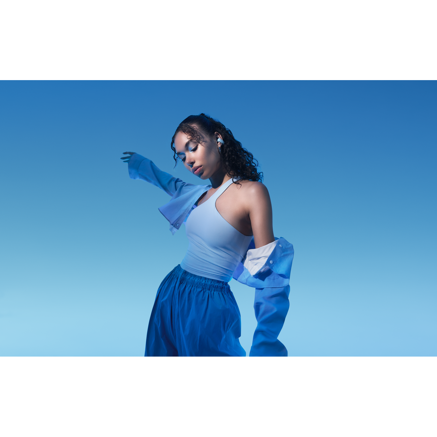 Een vrouw met oordopjes staat voor een blauwe achtergrond. Ze draagt een lichtblauwe top en jas, en een blauwe broek.