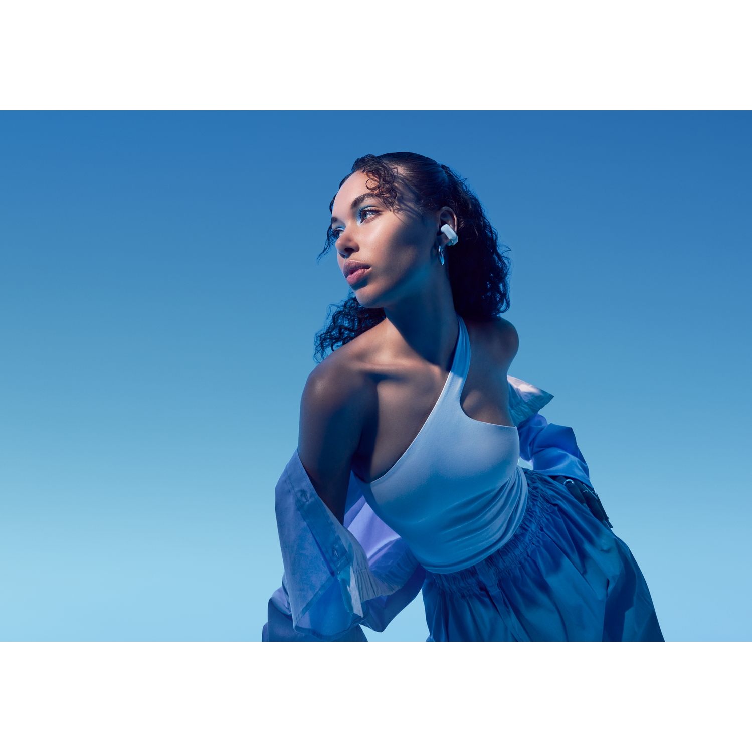 Een vrouw in een witte top en blauw overhemd met witte oordopjes, blauwe lucht.