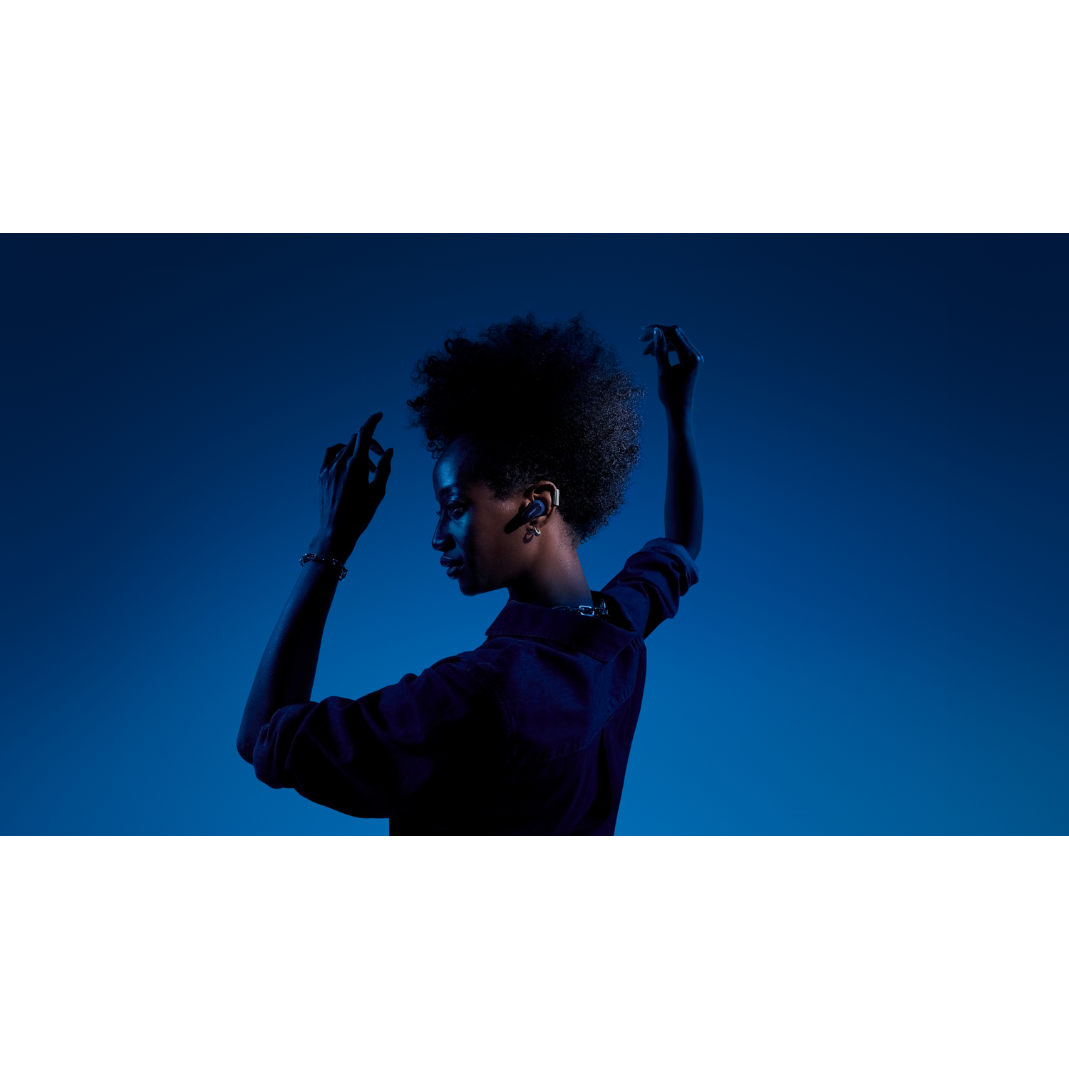 A woman with headphones, arms raised, in profile against a blue background, wearing dark clothing.