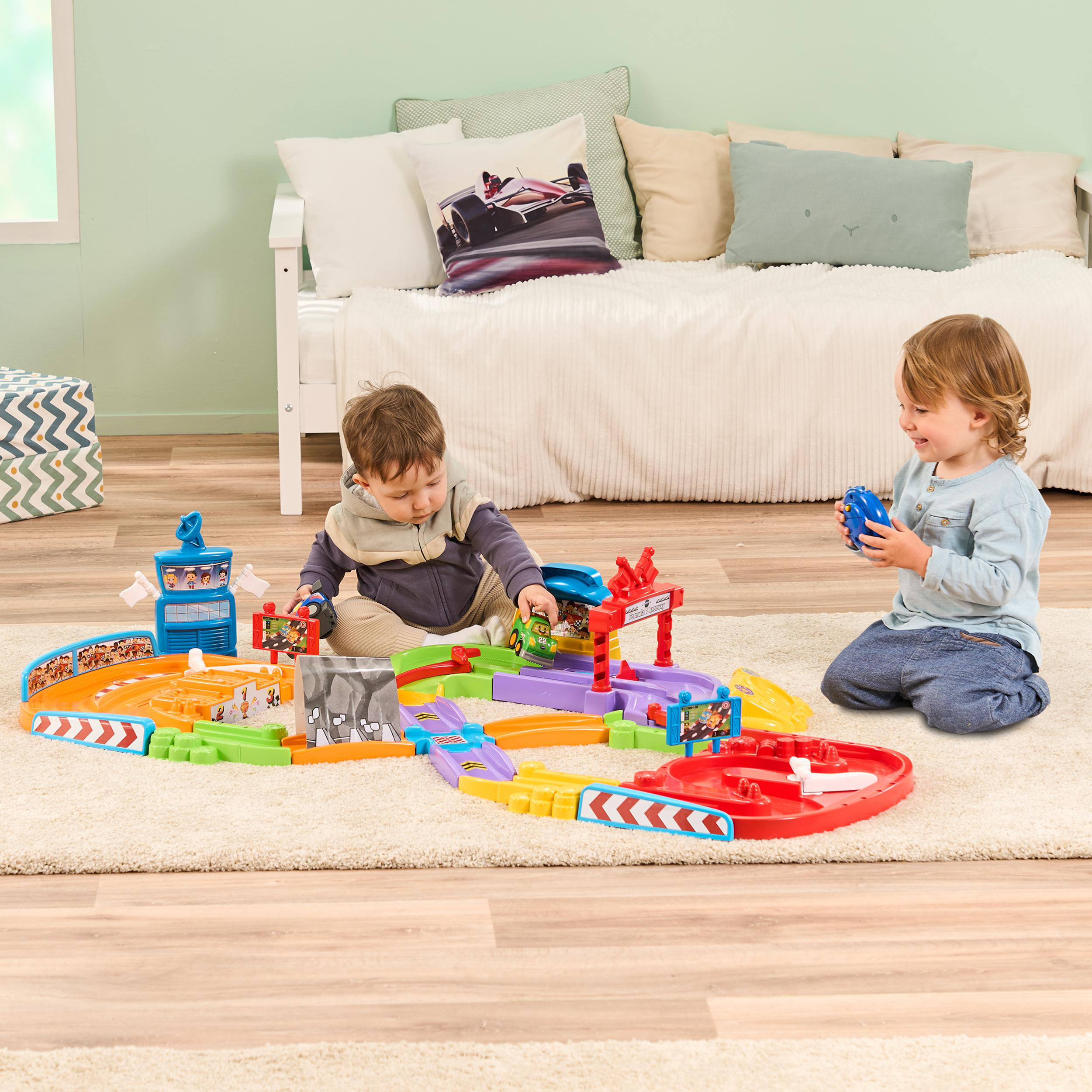 Zwei Kleinkinder spielen mit einer Spielzeug-Rennstrecke auf einem Teppichboden. Einer hält einen blauen Ball.