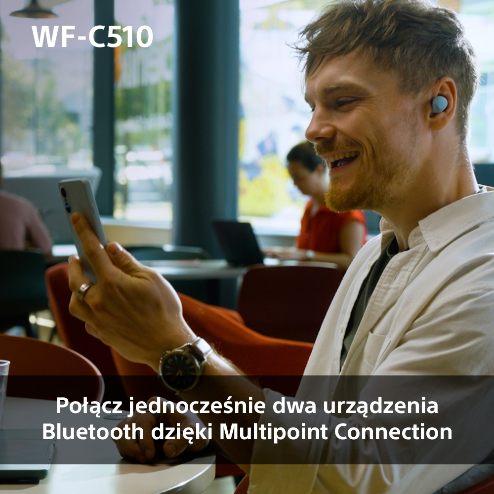 Mężczyzna używa słuchawek i smartfona z uśmiechniętą twarzą w kawiarni. Tekst: Połącz jednocześnie dwa urządzenia Bluetooth.